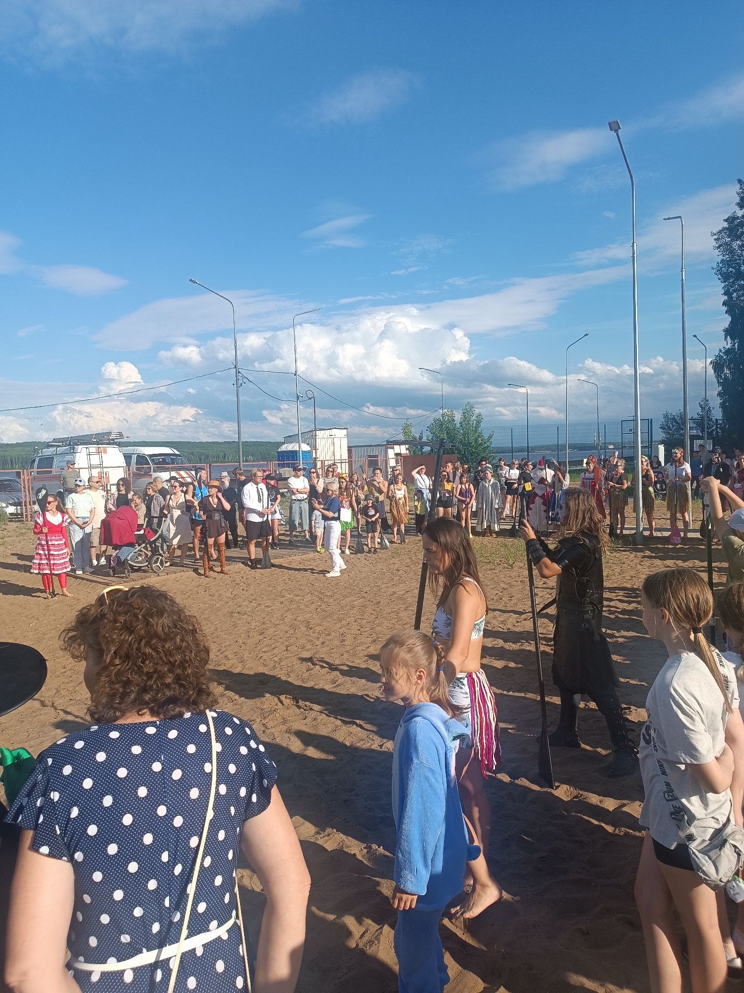 This media file shows a gathering of people on a sandy beach under a bright blue sky with scattered white clouds. The scene appears to be outdoors in daylight, likely late morning or early afternoon given the strong sunlight casting shadows. Several people are dressed in costumes, suggesting an event such as a festival, reenactment, or themed party.
In the foreground, a woman with curly hair wearing a dark blue dress with white polka dots is visible from the back. To her left, a child in a blue outfit is walking. Further into the scene, a group of people are standing and interacting. Some individuals are wearing costumes that include headdresses, loincloths, and what appear to be prop weapons. One person is dressed in a black leather-like outfit and holds a long staff. Another person wears a red and white patterned dress. Several individuals are dressed in casual summer attire.
In the background, there are vehicles, including vans, and structures that could be tents or temporary buildings. A fence runs along the edge of the gathering area. The presence of streetlights suggests this might be a public park or event space. The overall atmosphere seems relaxed and festive, with people mingling and observing the proceedings. The location appears to be near a body of water, indicated by the horizon line in the distance. No visible text is present in the image.