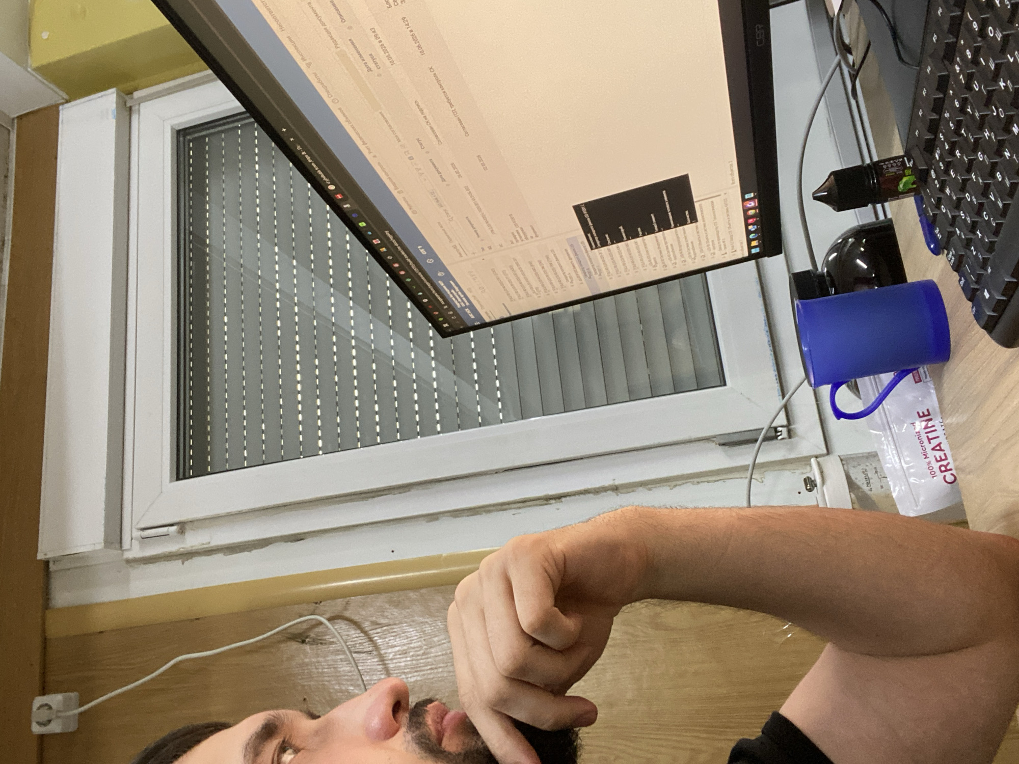 A medium shot captures a man from the chest up, seated at a desk and looking intently at a computer monitor. He is resting his chin on his right hand, his elbow supported by his forearm. His left arm is not visible. The man has dark hair and a beard. The computer screen displays a spreadsheet or database with text in what appears to be Russian. A blue mug, a dark bottle, and a small bottle with a green label are on the desk. A white package with the word "CREATINE" is also visible. The background shows a window with blinds, a wall, and a white electrical outlet with a wire. The scene is indoors, suggesting a home office or workspace. The overall lighting is dim, with a warm tone from an artificial light source.
