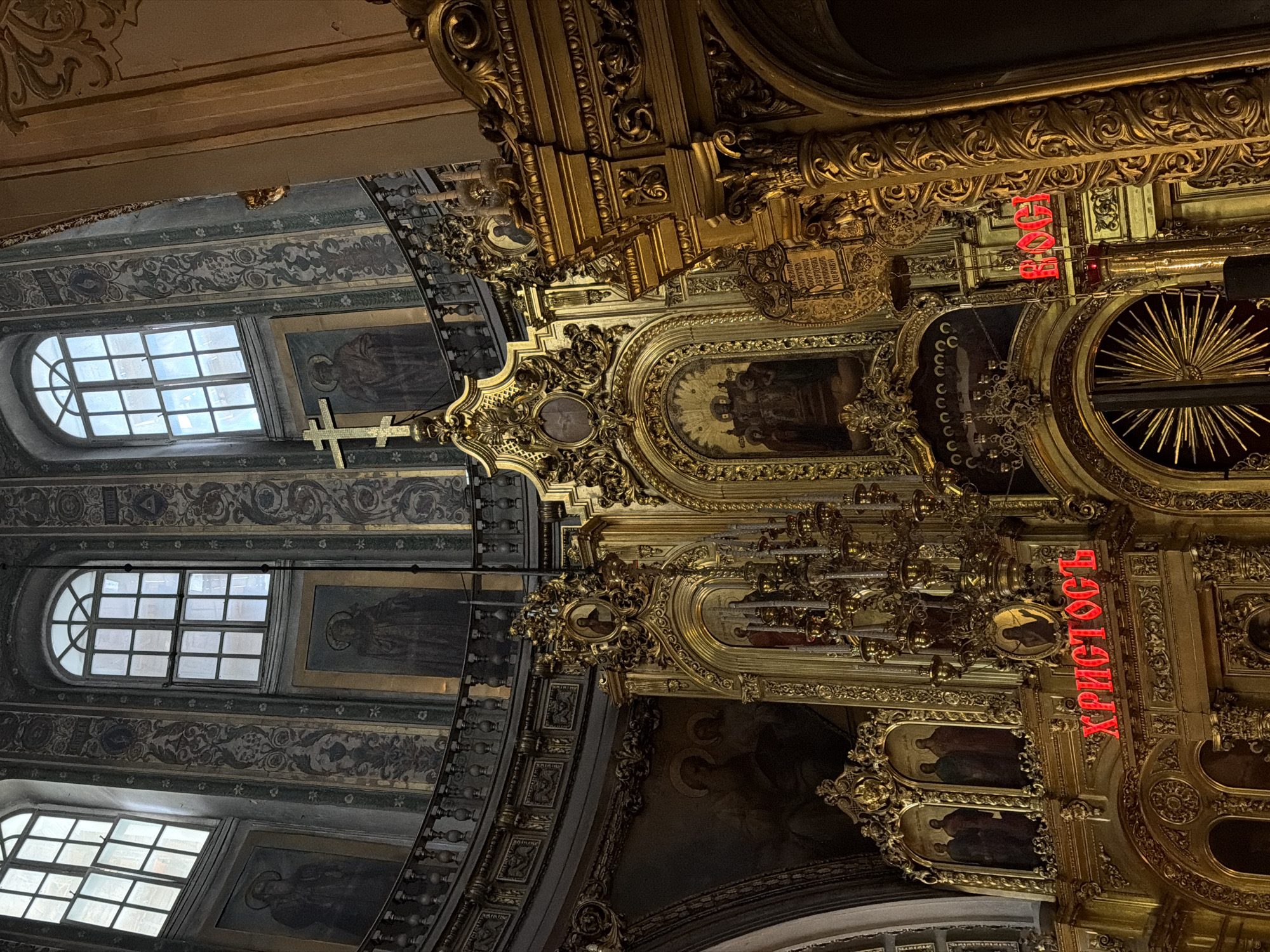 This is an interior view of a Russian Orthodox church in Moscow, Russia, likely a cathedral or major church due to the level of detail and ornamentation. The camera is tilted upwards, focusing on the ceiling and upper portions of the interior architecture.
Architectural Features: The building exhibits highly ornate gold decor, including detailed carvings and moldings. There are paintings integrated into the architecture, possibly icons or religious scenes. The walls are decorated with patterned wallpaper or painted designs. Windows are arched and divided into smaller panes.
Religious Elements: A cross is visible in the foreground. There are words written in Cyrillic script in red neon that say "Christ is risen," which is an Easter greeting.
Lighting and Atmosphere: The overall lighting is dim, but highlights the gold detailing. The image has a grand, religious atmosphere.