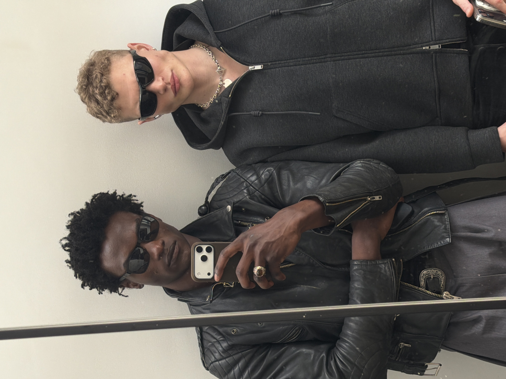 Two men are reflected in a mirror, possibly in London. The lower man is wearing a black leather jacket, dark sunglasses, and is holding an iPhone in his right hand, displaying the camera side towards the viewer. He has dark skin and short, curly black hair. He is wearing a ring. A decorative buckle is visible on the jacket. The man above him has fair skin, short blonde hair, and is wearing dark sunglasses and a gray hooded zip-up sweatshirt with a chain around his neck. He is holding a phone in his right hand. The reflection indicates they are in a room with plain, light-colored walls.