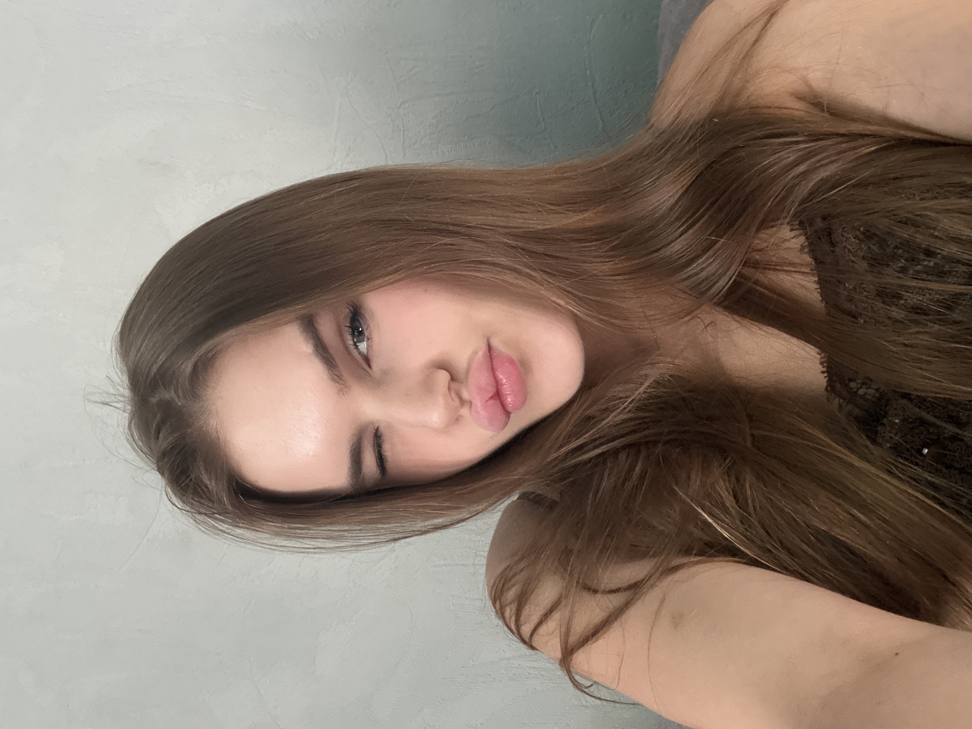The image is a close-up selfie of a young woman with long brown hair. She is winking one eye and puckering her lips in a kissing pose. Her skin is fair, and she appears to be wearing light makeup. She is wearing a dark-colored top with lace detailing. The background is a textured gray wall. The location is not specified in the image.