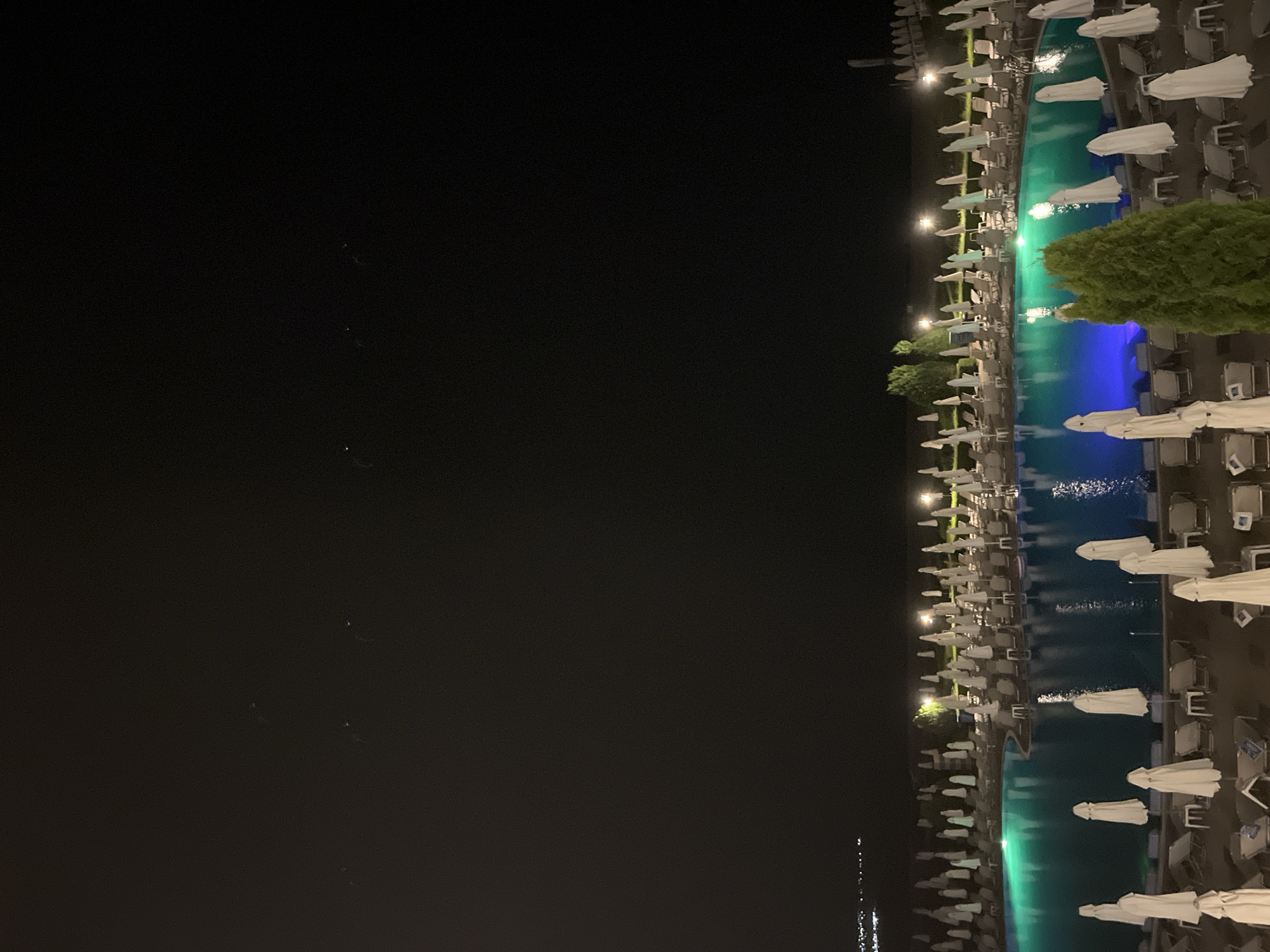 The image shows a night scene at a resort or hotel in Obzor, Bulgaria, with a swimming pool illuminated by green and blue lights. The pool is surrounded by rows of closed white umbrellas and lounge chairs. In the distance, the lights of the town of Obzor are visible along the coast, with a faint line of lights indicating structures and possibly a promenade. The sky is dark and clear, with a few small, indistinct bright spots that could be distant lights or atmospheric phenomena. The overall impression is one of a quiet, nighttime ambiance at a coastal resort.