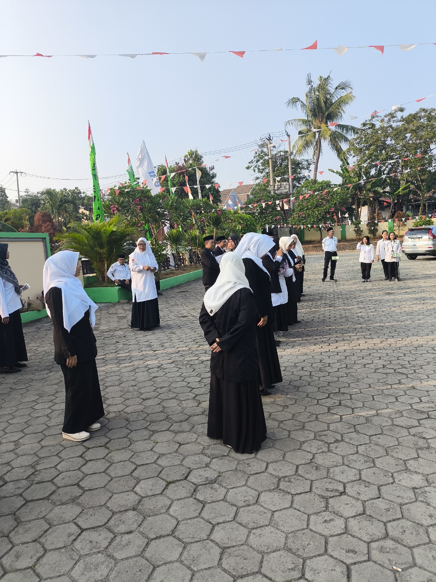 A group of individuals, predominantly women dressed in modest attire including headscarves and long skirts, are gathered outdoors. Many are wearing black and white outfits. The scene appears to be a school or public gathering, with flags and bunting decorating the area. The ground is paved with hexagonal stones. In the background, trees and buildings are visible, along with a white SUV. The sky is clear and blue, suggesting it is daytime. The location is identified as the yard of the State Junior High School 3 of Cilegon in Cilegon, Indonesia.