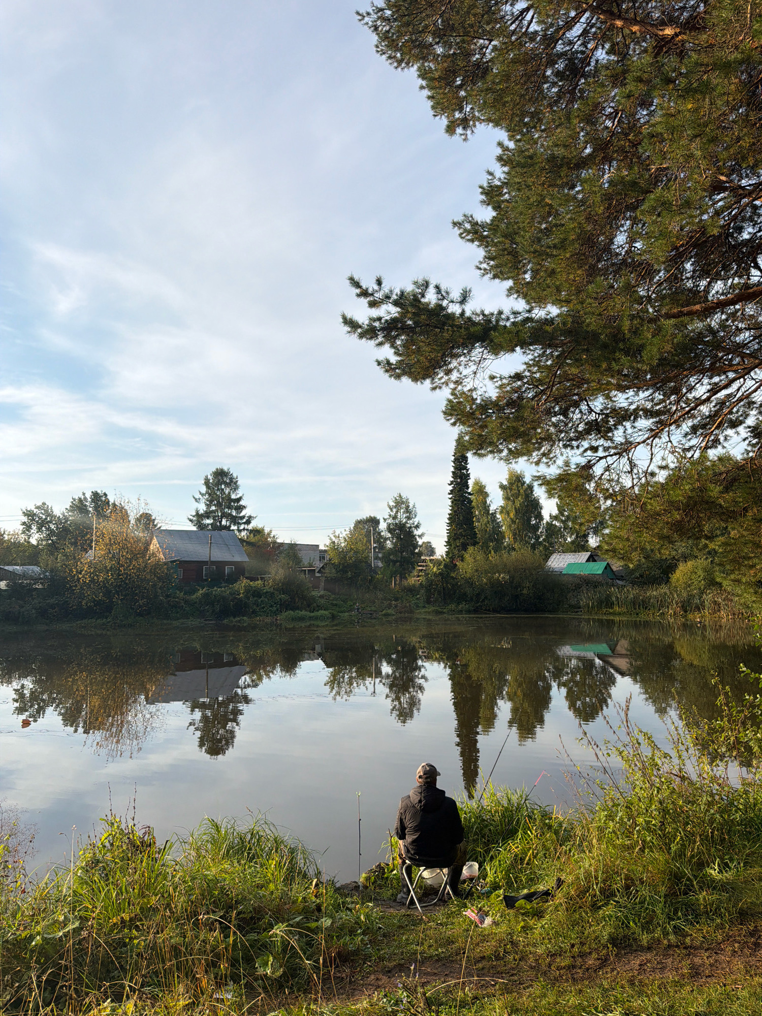 The image depicts a serene lake scene with a lone fisherman seated on a folding chair by the grassy shore. The fisherman, viewed from behind, is wearing a dark jacket and a baseball cap, facing the calm water. Two fishing rods are propped up in the water, suggesting an active fishing session. The surrounding vegetation is lush and green, with tall grasses and bushes bordering the water's edge. Across the lake, a small village is visible, with houses nestled amongst trees. The reflections of the houses and trees are clearly visible on the still surface of the water, creating a symmetrical and peaceful composition. The sky is partly cloudy with a soft blue hue, and the overall lighting suggests either early morning or late afternoon. The presence of a large pine tree in the foreground on the right adds depth and framing to the scene. The location is identified as Chaikovsky, Russia, a city known for its proximity to the Kama River and its natural landscapes.