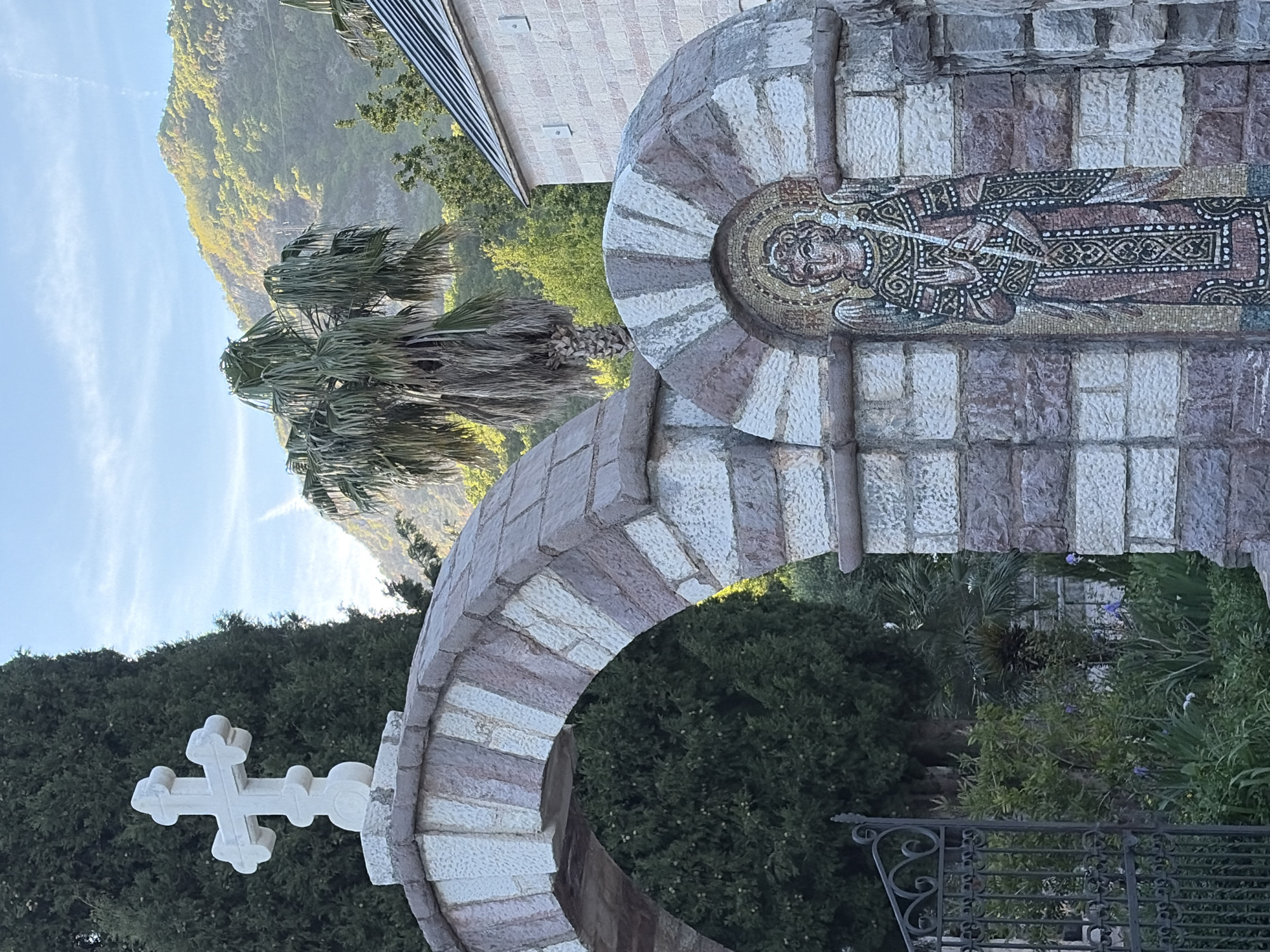 The image captures a serene outdoor scene in Budva, Montenegro, featuring architectural elements and natural landscapes under a clear sky. In the foreground, a prominent stone archway is visible, constructed from alternating light and darker-colored cut stones, creating a striped, radial pattern around its curve.
Embedded within the stonework above the arch is a vibrant mosaic depicting a stylized religious figure. The figure, possibly a saint, is rendered with intricate detail in small, colorful tesserae, wearing patterned robes and holding an object, possibly a staff or scroll. To the left of the arch, a white, ornate stone cross stands out against a backdrop of dense, dark green trees, possibly cypress.
Beyond the arch and trees, a majestic, verdant mountain rises into the distance under a bright blue sky, indicating clear weather. A large palm tree with long, feathery fronds is prominent in the middle ground, adding to the Mediterranean feel of the setting. Portions of a light-colored building with a patterned roof are visible in the upper right background, partially obscured by foliage. Beneath the arch, a lush garden area filled with various green plants and some small purple flowers can be seen, partially framed by a decorative black metal railing in the lower right.
The photograph appears to be taken during the daytime, likely morning or late afternoon, given the crisp light illuminating the scene. The overall atmosphere is one of historical tranquility and natural beauty, suggesting a religious site or monastery grounds. No explicit text is visible in the image.