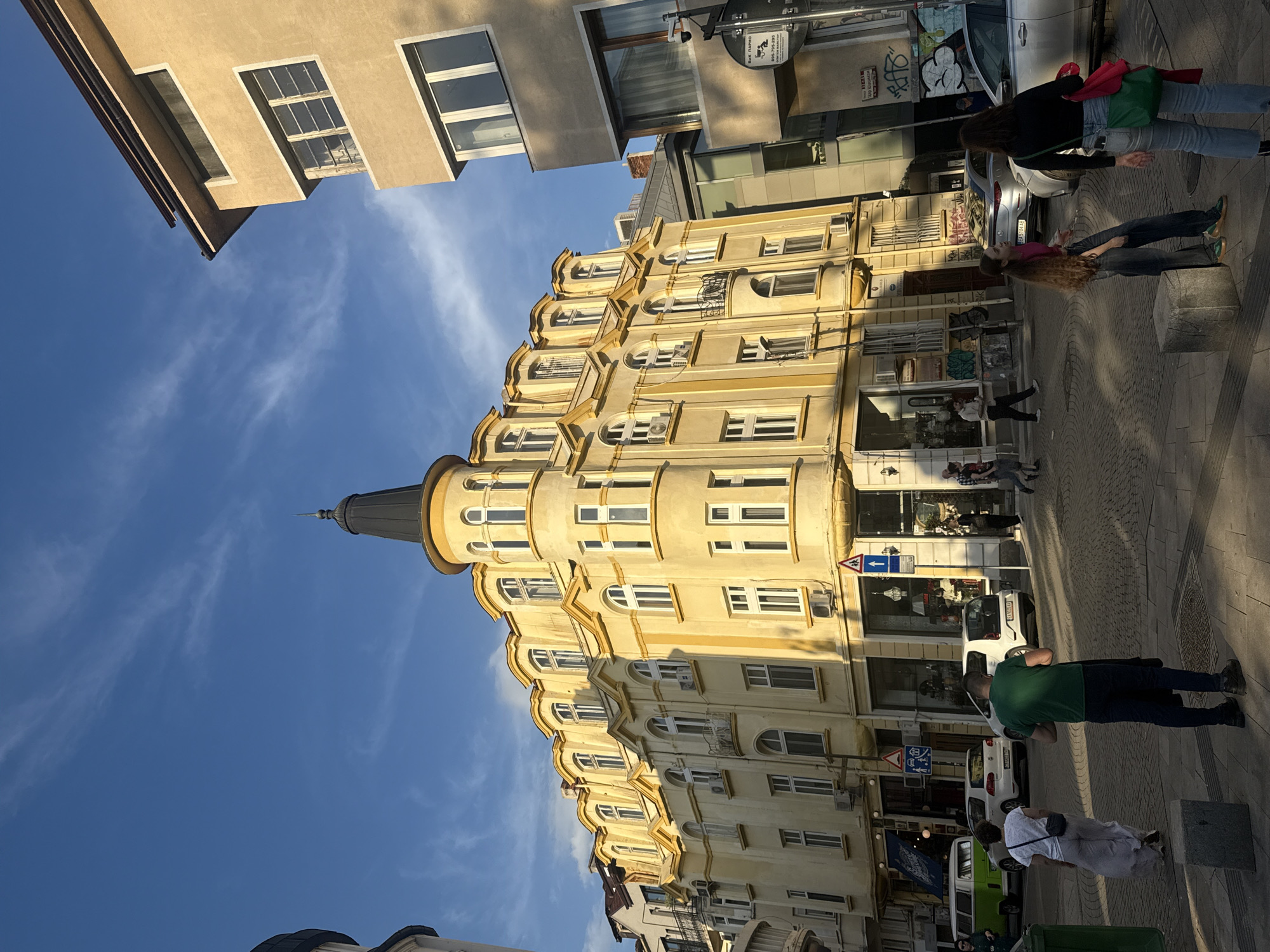The image captures a vibrant street scene, likely in Sofia, Bulgaria, characterized by an impressive, ornate building under a clear blue sky. The main subject is a large, light-yellow multi-story building featuring classical architectural elements, such as arched windows, decorative cornices, and a prominent round tower with a dark conical roof on the left side. Sunlight, seemingly from a low angle (either morning or late afternoon), illuminates the building, casting long shadows across the street and sidewalk.
Several people are present on the sidewalk, engaging in casual street activities. In the foreground-right, a person wearing a green t-shirt and dark pants stands, looking down. Further back, another individual in a white dress is seen from behind. In the middle ground, a person with long, light brown hair and a pink top stands near a grey concrete block, facing towards the street. Behind them, another person in a black jacket and jeans, carrying red and green bags, also stands on the sidewalk. Other indistinct figures are visible further down the street near shop entrances.
The street level features several shopfronts with large glass windows, reflecting parts of the opposite buildings. Multiple vehicles are parked along the street, including a silver sedan, white cars, and a green van. Visible street furniture includes traffic signs, such as a white arrow pointing left and a blue 'no parking' sign. A circular sign affixed to a building clearly shows "БИМЕ ТАПРО" and a phone number "0896-790-399". Graffiti is also present on a wall behind a parked car. The overall impression is a bustling urban environment bathed in natural light, showcasing both architectural heritage and daily city life.