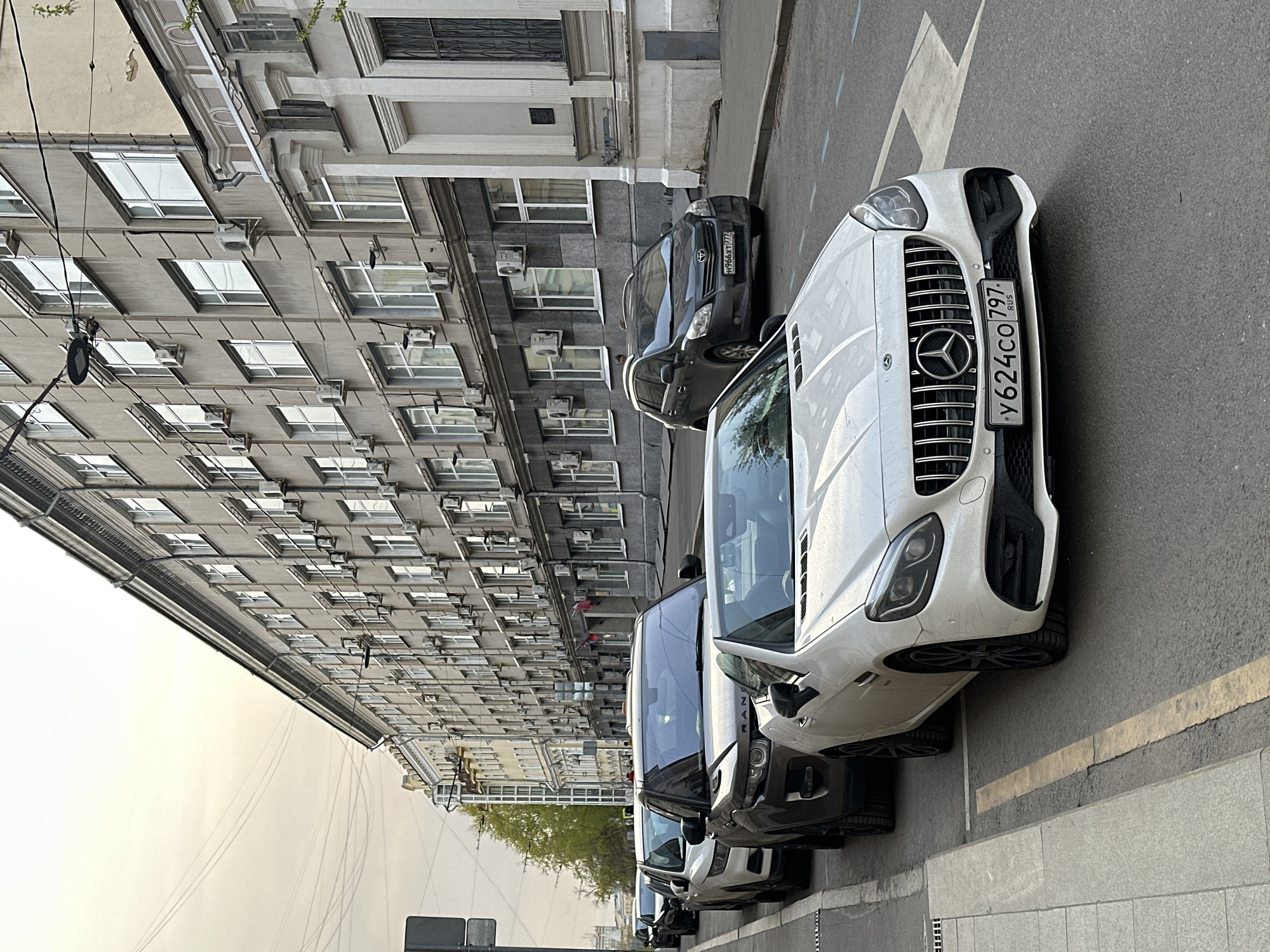 The image shows a street scene in Moscow, Russia. The foreground features a row of parked cars, including a white Mercedes-Benz AMG with license plate number "У624СО 797 RUS". To the left of the Mercedes, there are several other vehicles of differing colors. Behind the cars is a multi-story building with a gray facade, numerous windows, and external air conditioning units. The sky appears hazy or overcast. The building suggests a dense urban environment. The timestamp is not available. No people are visible.