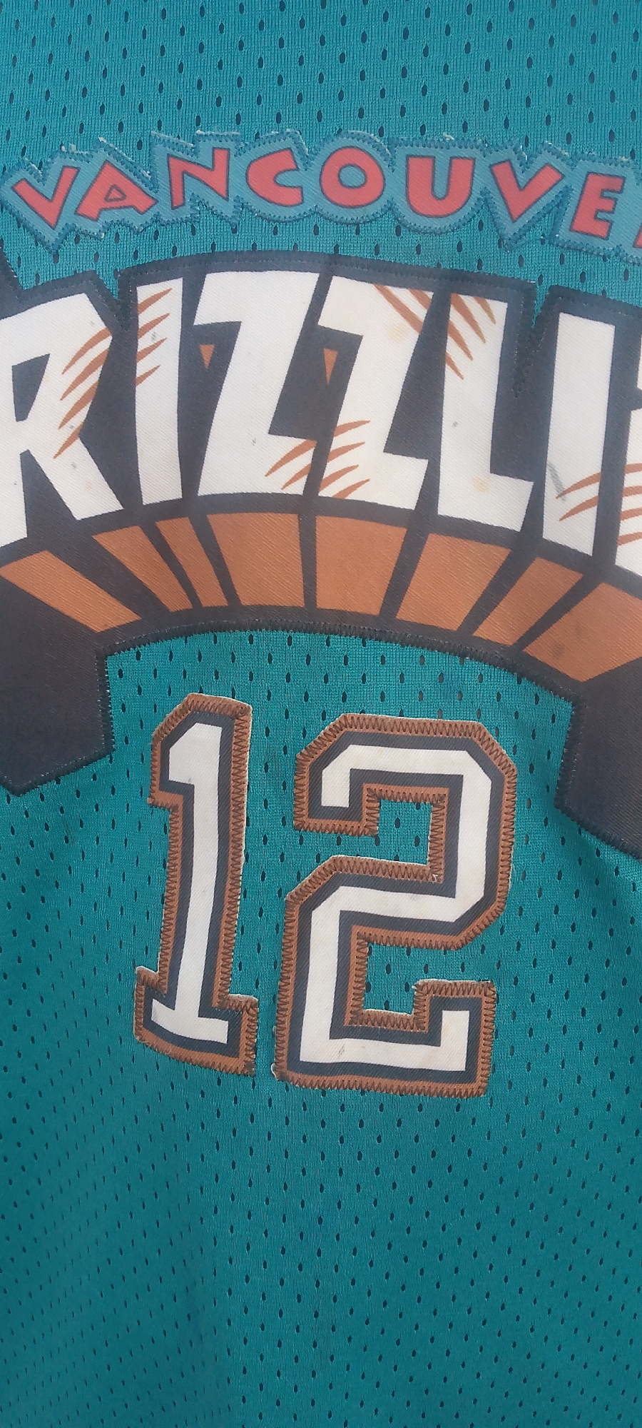 The image is a close-up, vertical shot of a teal-colored mesh jersey. The jersey features the word "VANCOUVER" in red, blocky lettering with a light blue outline and a jagged edge, positioned at the top. Below this, the word "GRIZZLI" is visible in large, white, distressed block letters with orange and black accents. The jersey also displays the number "12" in prominent white digits, outlined in black and orange. The material of the jersey is a fine mesh with a subtle pattern of small, uniform holes. The overall lighting is even, with no clear indication of the time of day or weather conditions. The image does not contain any people or indicate any specific urban or natural environment. It is solely focused on the details of the garment. There is no discernible connection to the city of Jalingo, Nigeria.