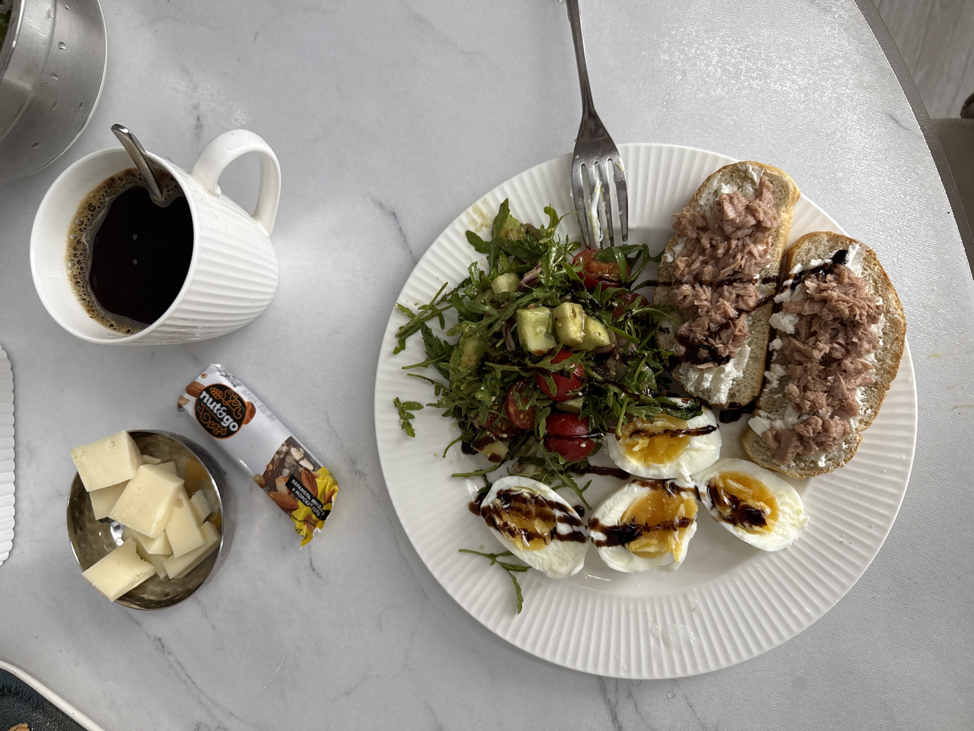 A high-angle, medium shot captures a breakfast spread on a white, marble-patterned table. In the upper left, a small, silver, textured bowl contains several cubes of light yellow cheese. Next to the bowl, a white and gold wrapper with the word "nutrigoo" is partially visible. In the upper right, a white ceramic mug with vertical ridges is filled with dark coffee, and a silver spoon rests inside. The main subject is a white, fluted plate in the center of the frame, laden with food. Three halved hard-boiled eggs, drizzled with a dark sauce, are positioned on the left side of the plate. To their right, a salad composed of mixed greens, cherry tomatoes, diced cucumber, and possibly avocado is visible. In the lower right portion of the plate are two open-faced sandwiches, each topped with a white spread, likely cream cheese, and shredded tuna. A fork rests on the edge of the plate. The background consists of the white, marble-patterned table. The scene depicts a breakfast setting, likely in a home environment in Nizhnyayarkeyevo, Russia, given the context. There are no people visible in the image.