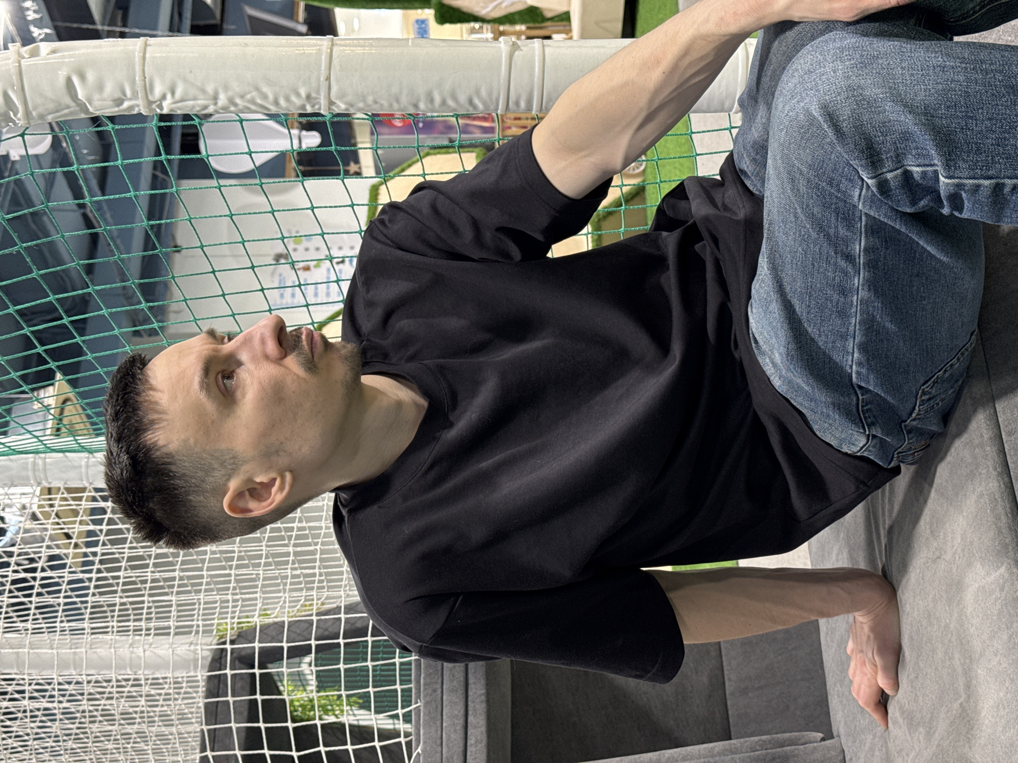 An indoor, vertical shot captures a man in a recreational setting, identified as Izhevsk, Russia. The subject, a man in his 20s or 30s with short dark hair, a light mustache, and stubble, is positioned sitting or leaning on a gray upholstered surface. He wears a plain black short-sleeved t-shirt and light blue denim jeans. His right arm is extended behind him, supporting his weight on the gray surface, while his left hand rests near his bent left knee. He is looking upwards and slightly to his right, with a neutral, possibly thoughtful, expression.
The background is dominated by green and white safety netting, indicating an enclosed play area, sports cage, or similar facility. Above the netting, white padded rails are visible. Through the mesh, glimpses of artificial green turf and other structures can be seen. The lighting suggests a well-lit indoor environment. No text is visible in the image. The scene conveys a moment of rest or observation within an active, indoor recreational space.