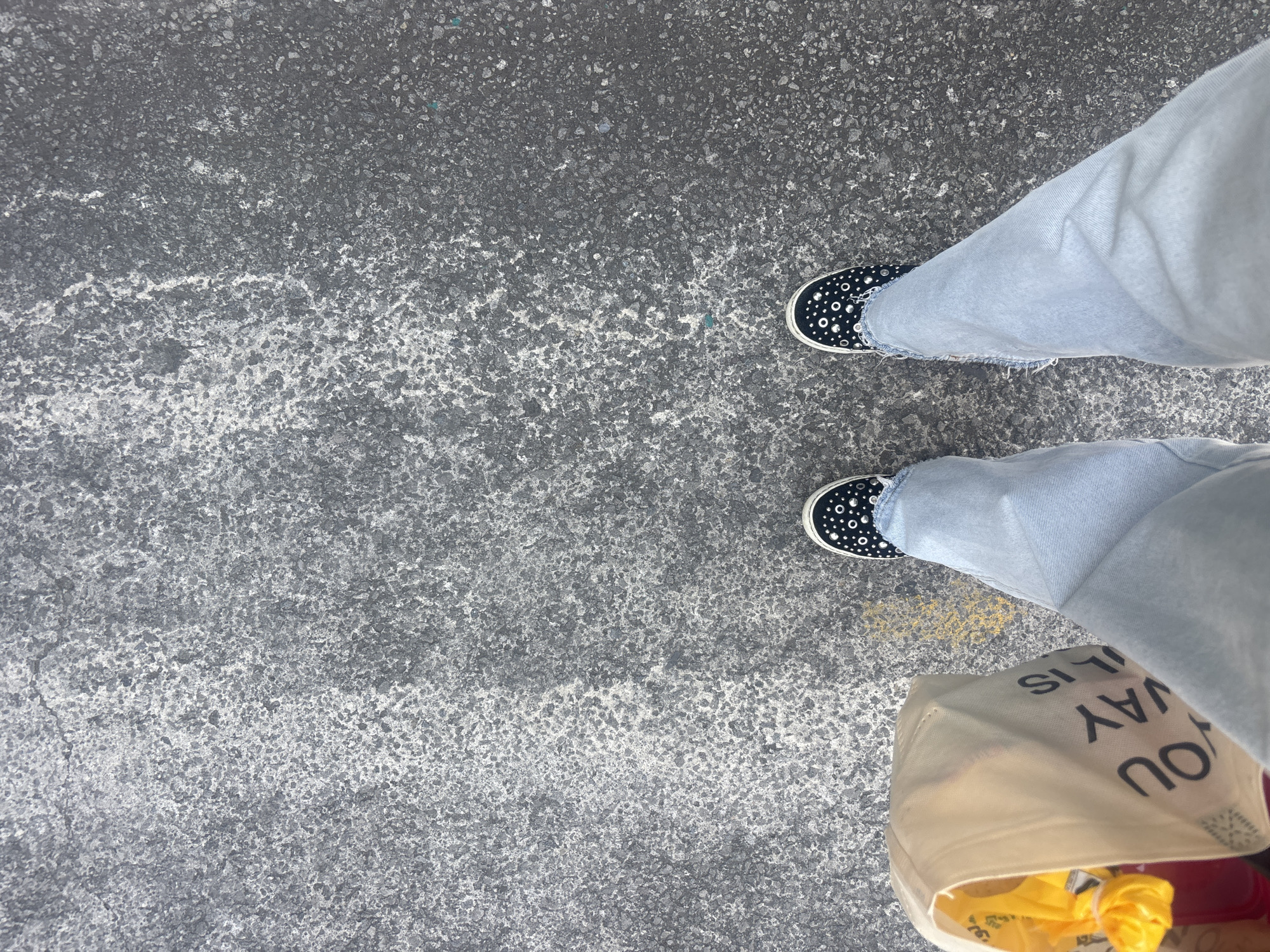 An overhead shot captures a person's lower legs and feet on a textured asphalt or concrete surface in Buenavista, Mexico. The person wears light blue, possibly denim, jeans with frayed hems. On their feet are black slip-on shoes adorned with numerous small, silver-colored studs.
The ground is predominantly dark grey with lighter grey and white speckled patches, indicating wear or possibly dried residue. A faded streak of yellow paint is visible on the pavement near the right shoe. The lighting is bright and even, suggesting daytime.
The person is holding a light-colored, possibly beige or off-white, cloth tote bag, visible in the lower right corner of the frame. The bag contains items, including something yellow and possibly red. Partial, inverted text is visible on the bag, reading "YOU WAY IS". The activity depicted is standing or walking on this paved surface, likely a street or sidewalk. The image focuses on the lower body, making emotions undiscernible.