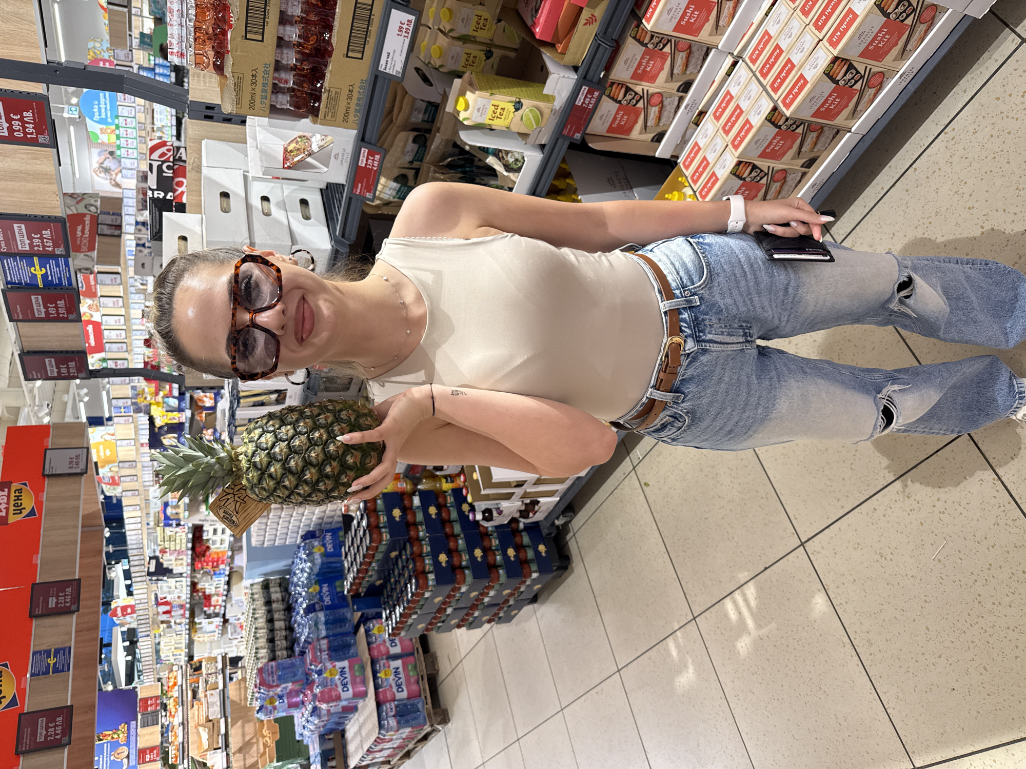 A woman is holding a pineapple in a supermarket aisle. She is wearing sunglasses, a beige tank top, ripped blue jeans, and a watch. She is standing next to shelves stocked with various food products, including drinks, sushi kits, and other packaged goods. Price labels in Bulgarian Levs and Euros are visible on the shelves. The scene appears to be inside a LIDL supermarket, likely in Blagoevgrad, Bulgaria, based on the local currency displayed.
