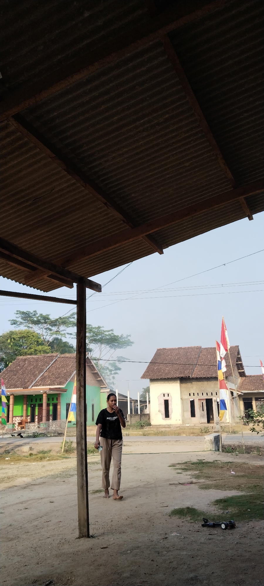 The image depicts a somewhat rural outdoor scene, possibly in the village of Tanjung Ratu Ilir, Indonesia, under a clear sky. In the foreground, a corrugated metal roof structure supported by wooden beams casts a shadow over the scene. A young woman, wearing a black t-shirt and light brown trousers, walks from left to right. She appears to be holding something to her mouth, possibly food or a cigarette, and carries a light-colored object in her left hand. Her feet are bare.
Behind her, the environment consists of dusty ground with patches of dry grass and a few scattered objects, including a black toy car with white wheels in the lower right corner. Two houses are visible in the mid-ground. The house on the left is painted green with a brown tiled roof and has several windows and a porch. Several flags, including what appear to be Indonesian flags (red and white), are displayed in front of this house. The house on the right is a simpler structure with a brown tiled roof and light-colored walls, featuring two large, dark rectangular openings that suggest unfinished windows or doorways. A few more flags are also visible in front of this house. Trees with green foliage are scattered throughout the background, and power lines are visible in the upper sky. The overall atmosphere suggests a casual, everyday setting.