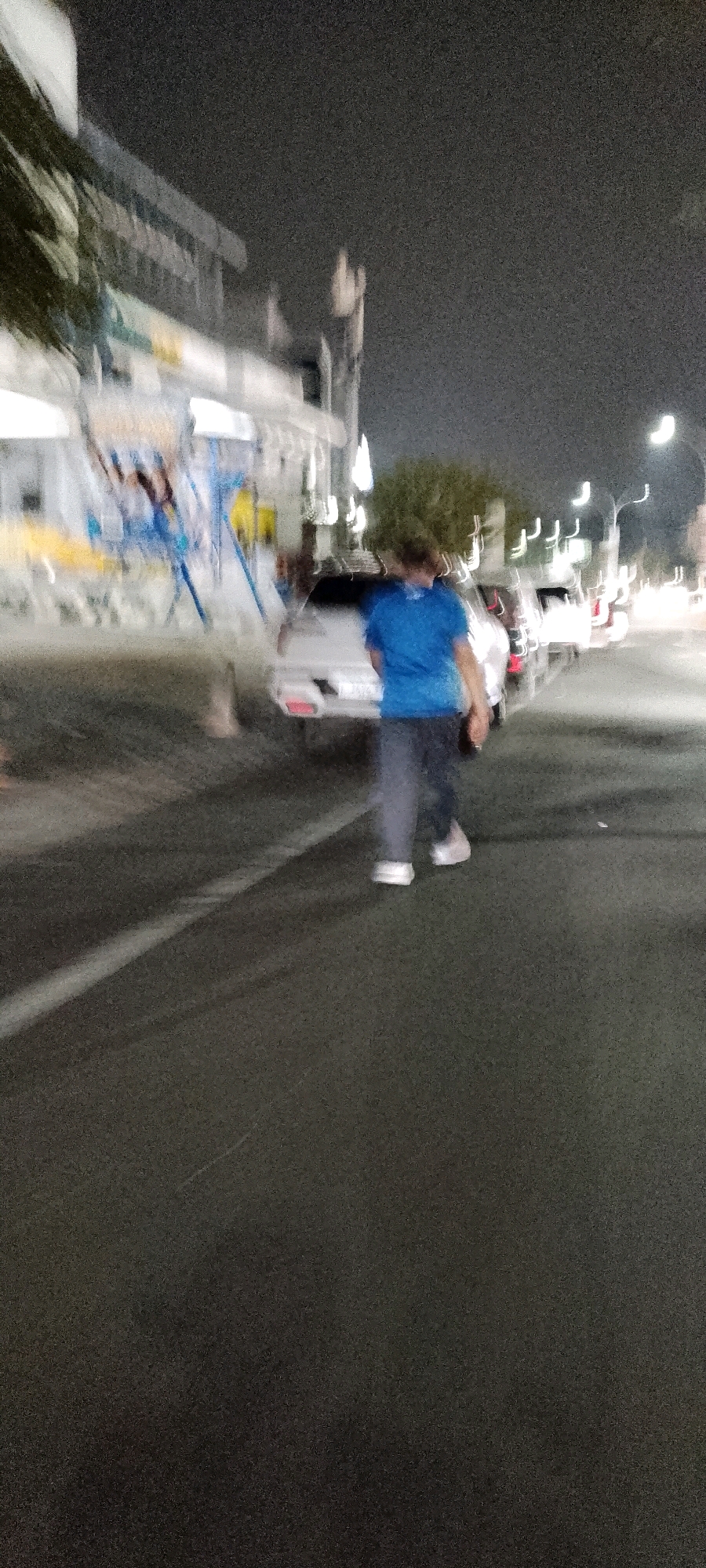 A blurry, night-time photograph taken on a street in Gunung Putri, Indonesia, shows a person walking on the asphalt road. The individual, wearing a blue t-shirt and dark pants, is in motion, as indicated by the blur. Several cars are parked along the side of the road, with their headlights and taillights illuminated, suggesting the presence of street lighting and possibly a commercial area. The background features buildings and trees, also rendered indistinct by the low light conditions and motion blur. A white line runs along the road, dividing lanes or marking a pedestrian walkway.