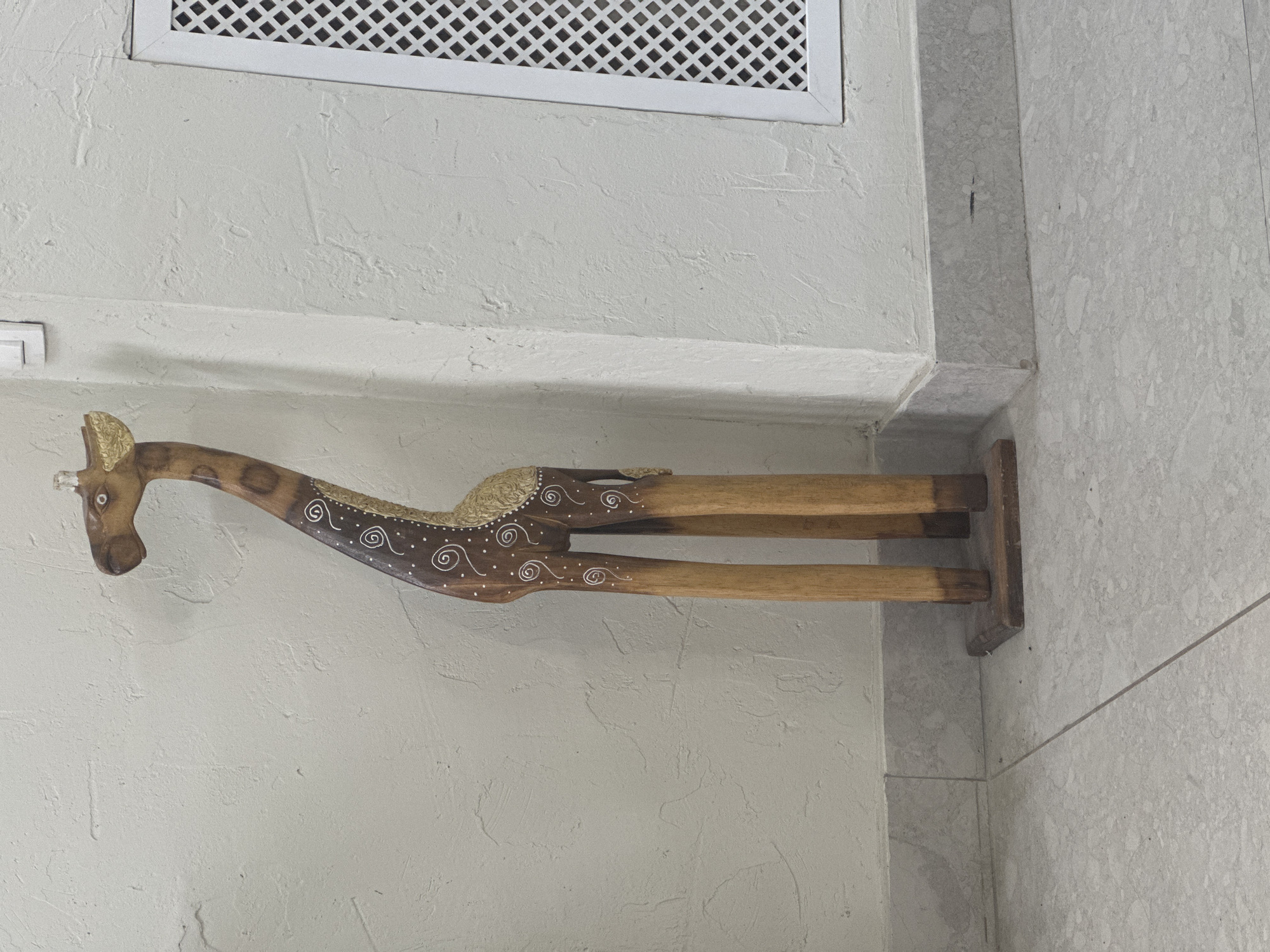 A wooden giraffe figurine stands on a tiled floor against a textured white wall. The giraffe is tall and slender, with a wooden base and four wooden legs. Its body is brown with decorative white swirls and gold accents. A white vent is visible on the right side of the wall. The scene appears to be indoors.