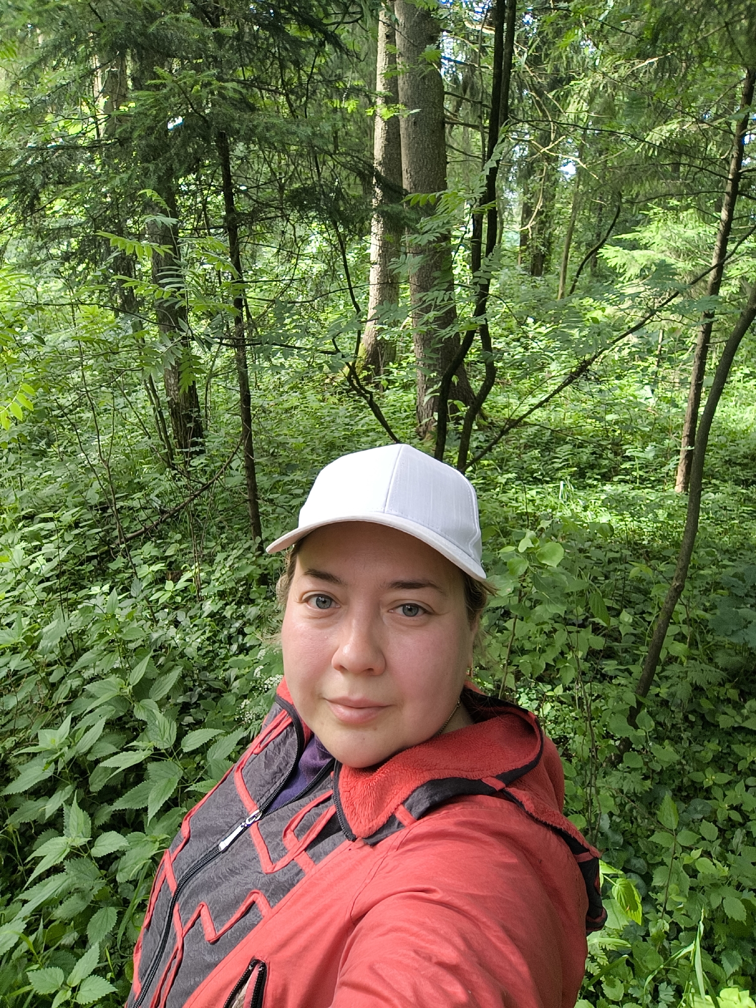 A selfie is taken in a dense forest. A person wearing a white baseball cap and a red jacket with black accents is visible from the chest up. The person's face is turned towards the camera, and they have fair skin and blue eyes. The background is filled with lush green foliage, including trees, bushes, and undergrowth. The lighting suggests it is daytime, with sunlight filtering through the leaves. The location is described as Novonikolskoye, Russia.