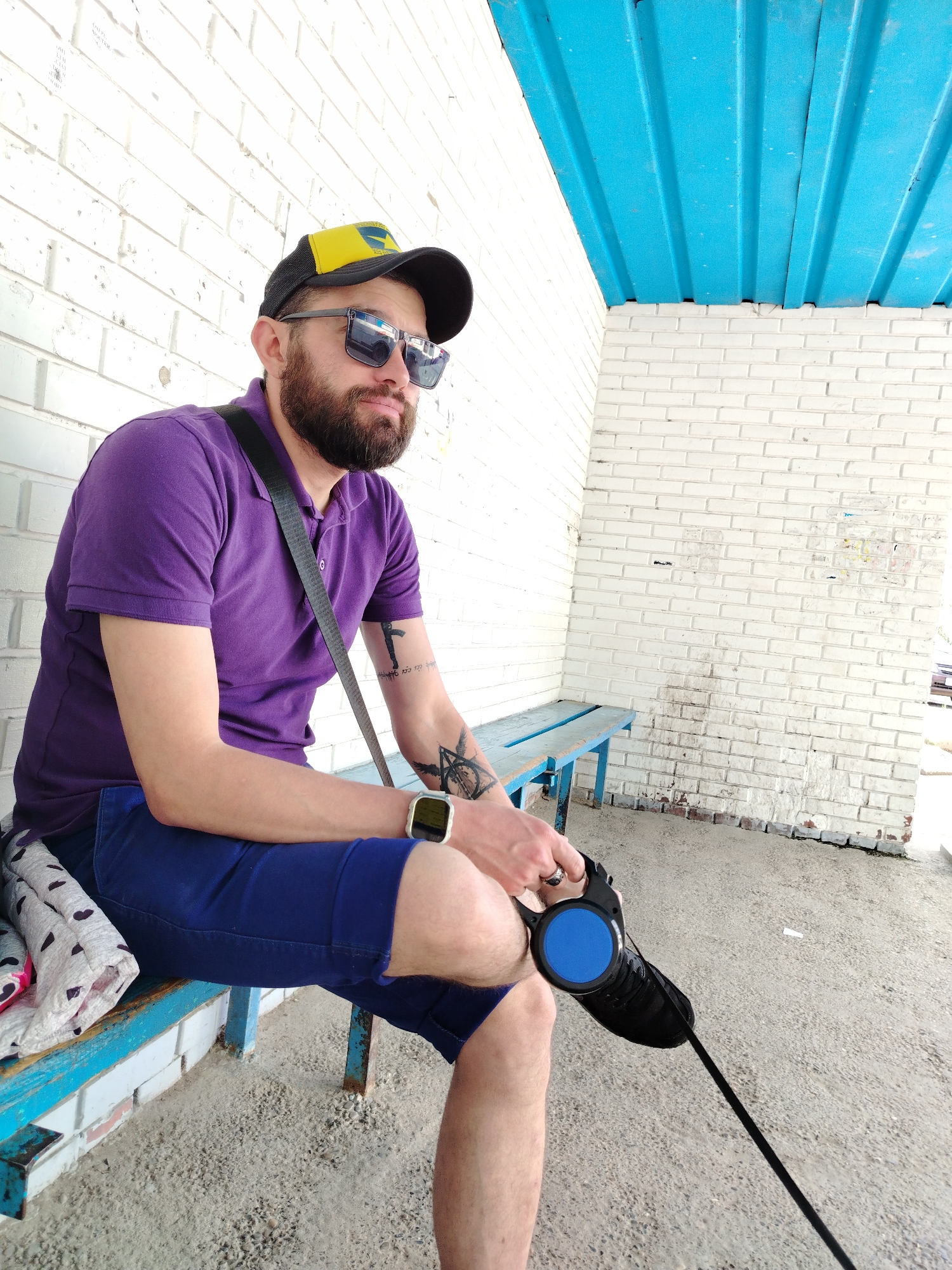 A single adult male sits on a worn, blue-painted bench in Noviy, Russia. He has a full beard and wears a black and yellow baseball cap, square-framed sunglasses, a purple polo shirt, and blue shorts. A dark strap crosses his torso. Tattoos are visible on both forearms: a stylized rifle with text on the right, and a geometric emblem on the left. He wears a gold-colored smartwatch on his left wrist and a ring on the fourth finger of his left hand. His left hand grasps a black retractable dog leash with a prominent blue circular button. A black shoe is partially visible near the leash mechanism. On the bench beside him, a folded white fabric with dark heart patterns is visible. The bench is positioned against a white brick wall showing extensive wear, dirt streaks, and residue from old postings. Above, a light blue corrugated metal roof provides shelter. The ground surface is rough concrete.