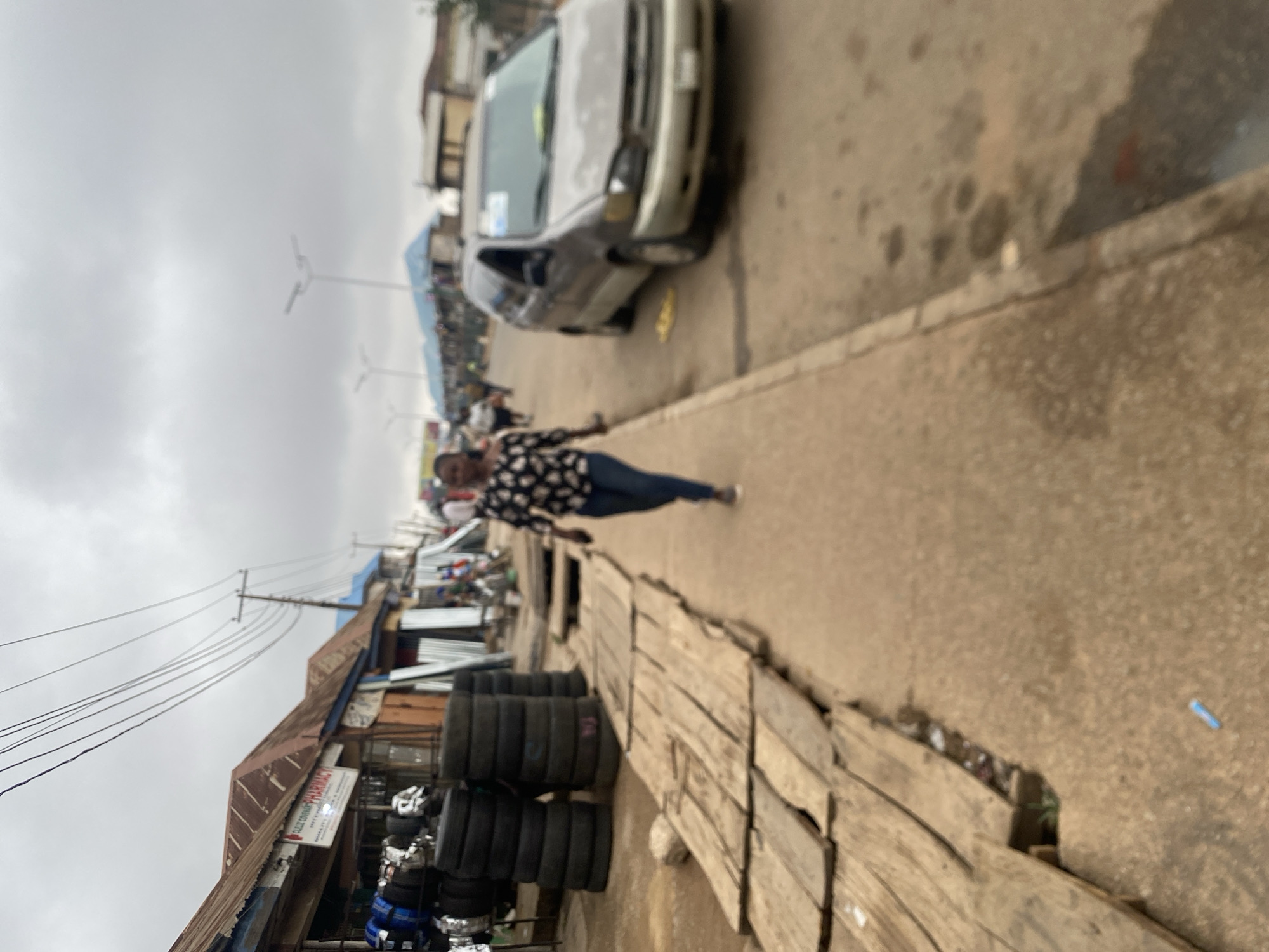 The scene is a street in Bukuru, Nigeria. A woman in a patterned top and blue jeans is walking along the sidewalk. To her left, there is a stack of tires and a shop. Behind the shop, power lines are visible. A silver car is parked on the street, partially blocking the view down the road where more people are visible. The sky is overcast.