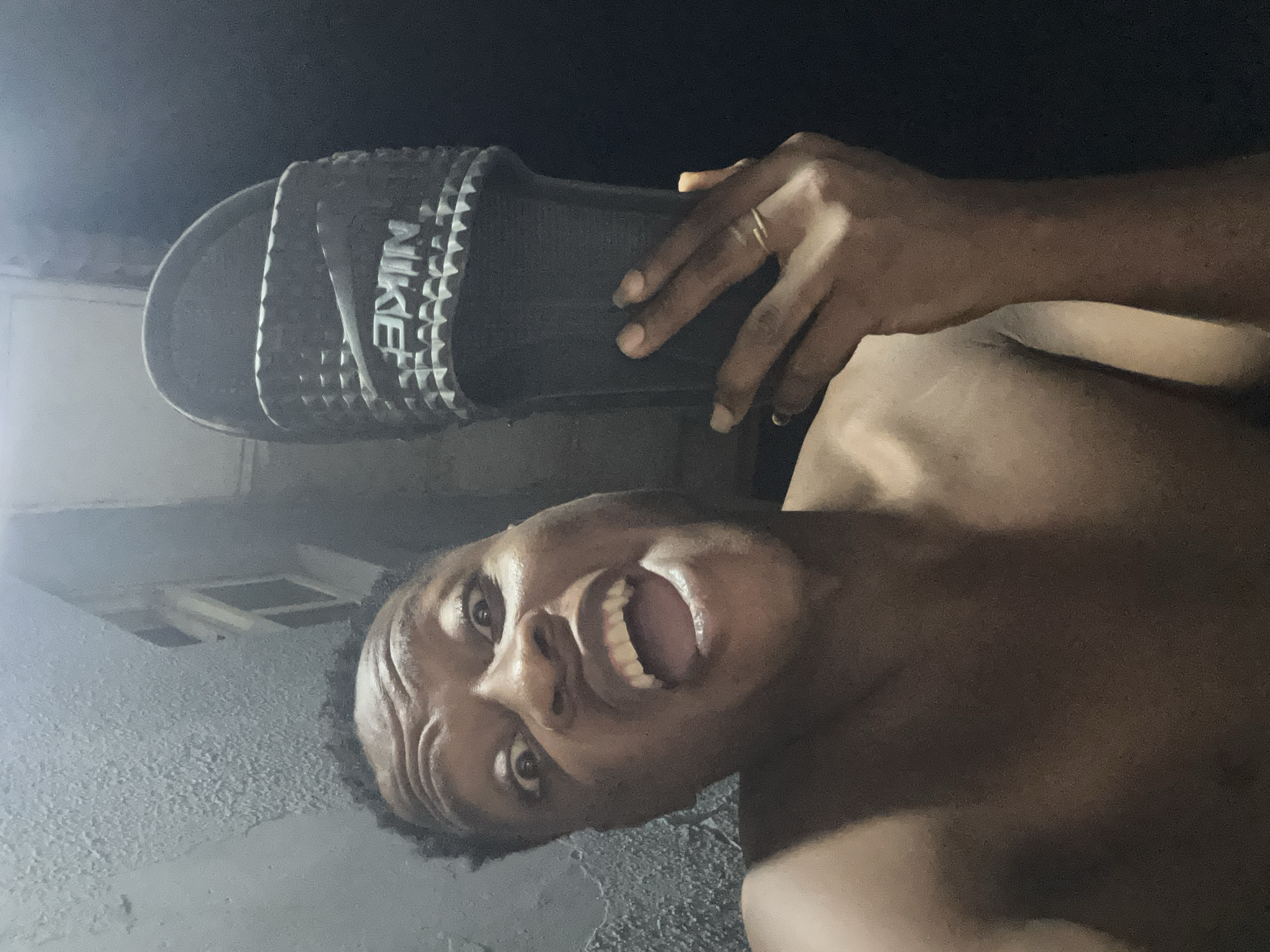 An adult male individual is depicted in a close-up, self-portrait style photograph taken in the city of Ikorodu, Nigeria. The subject is shirtless and has a dramatic facial expression with wide eyes, raised eyebrows displaying prominent wrinkles, and an open mouth revealing upper and lower teeth. His right hand is raised, holding a black Nike brand slide/slipper which features a textured strap embossed with the Nike logo and swoosh. A thin, gold-colored ring is worn on the ring finger of his right hand. The background on the left consists of a textured, light-colored wall, while the upper right background reveals a partial view of a building with a window, obscured by dim lighting. The overall illumination of the scene is low.
