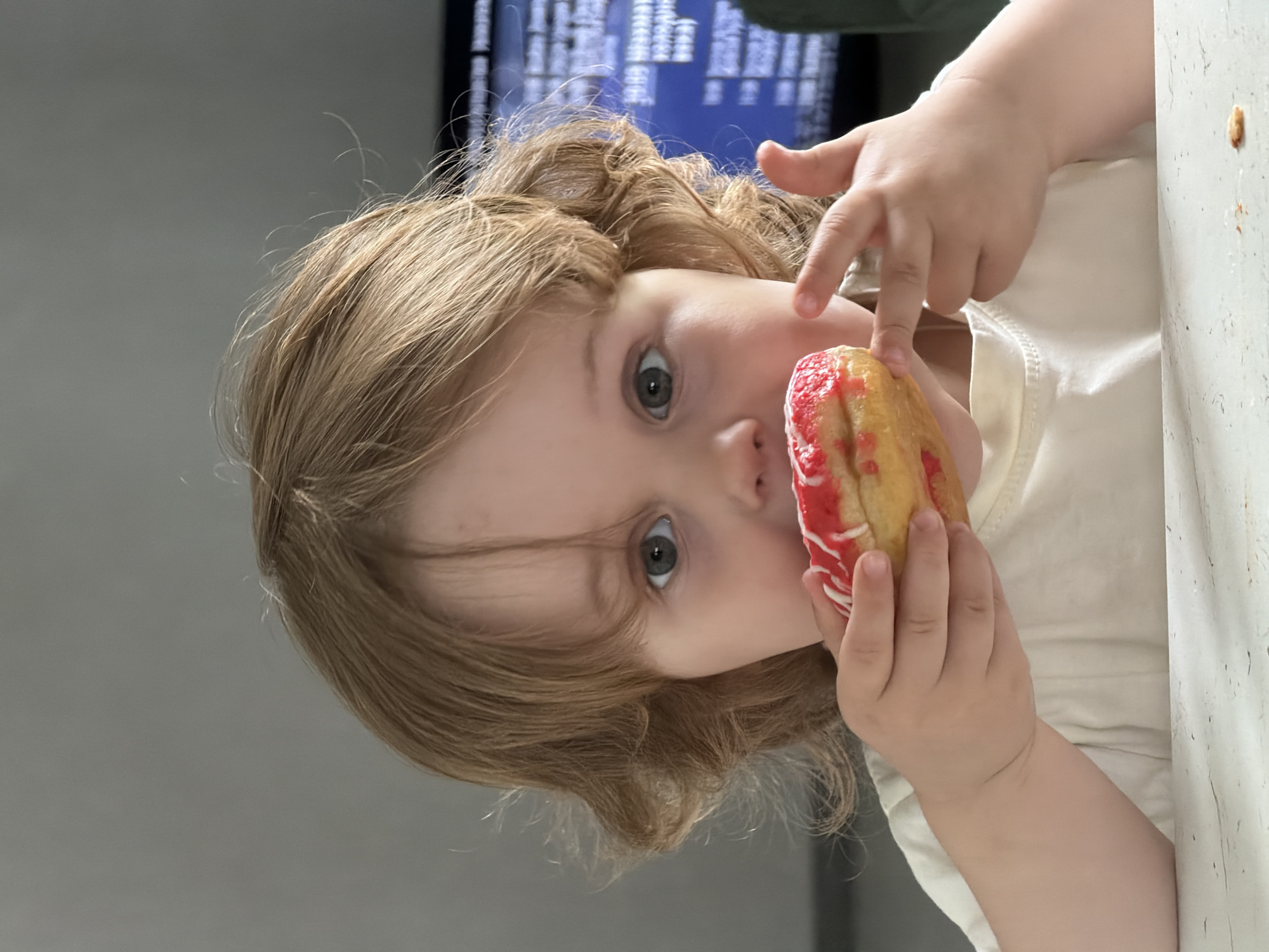 A young child with light brown hair is holding a donut with red icing and white stripes and bringing it to their mouth. The child is wearing a white t-shirt. The background shows a wall and a blurred television screen displaying text. The scene appears to be indoors, likely in a home setting, possibly in the city of Kamyennoye, Russia. There is a white table in front of the child.