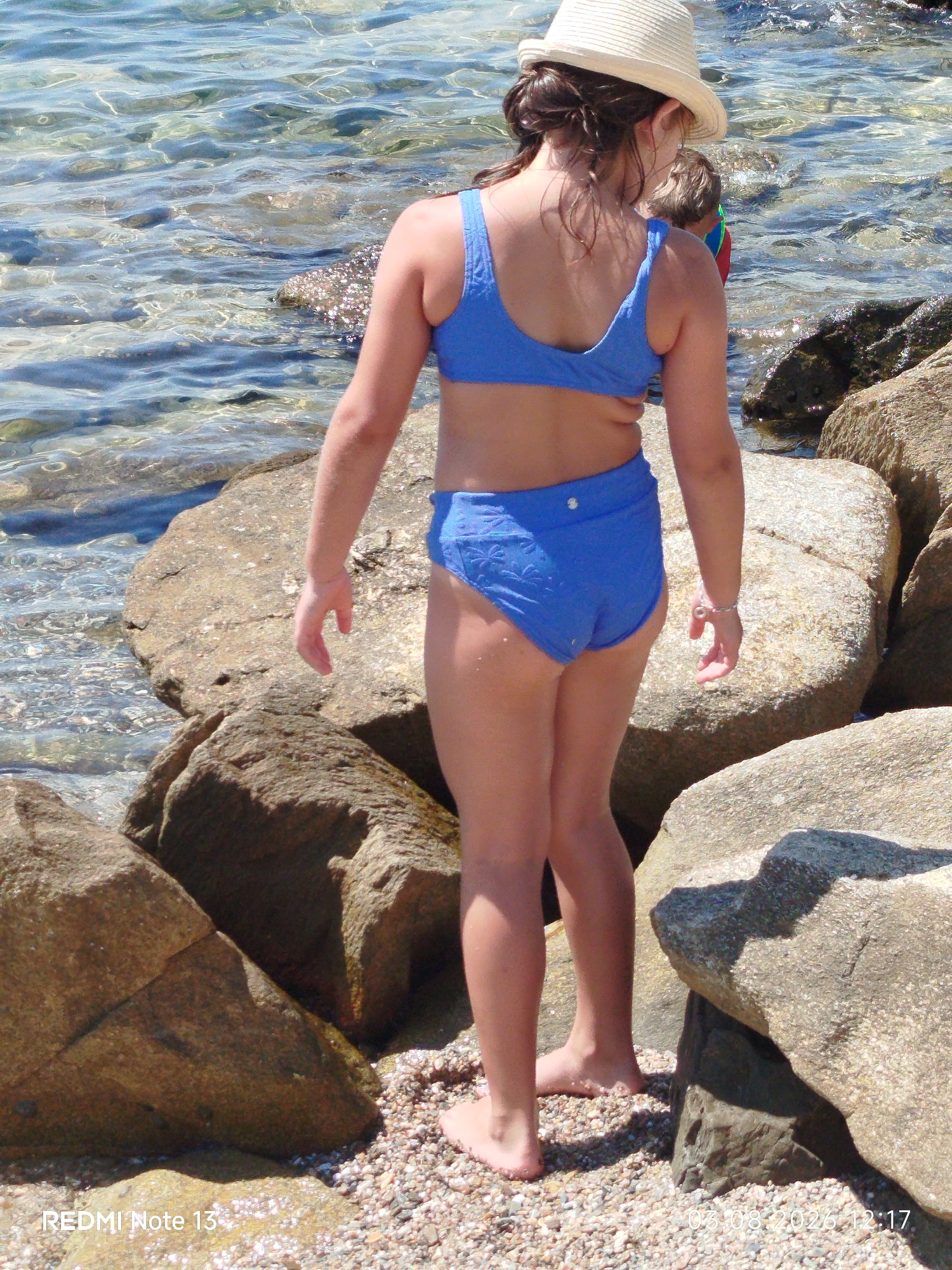 This is a photograph of a young girl wearing a blue bikini and a straw hat, standing on a rocky shore by the sea. She is facing away from the camera, looking out at the water. The water is clear and shallow, with sunlight glinting off its surface. The rocks are large and rough, with some pebbles and sand scattered around them. In the background, another child can be seen partially submerged in the water, wearing a colorful swim shirt. The sun appears to be shining brightly, suggesting a warm, sunny day. The overall scene evokes a sense of leisure and enjoyment of a beach environment. The image has a timestamp at the bottom right corner which reads "03.08.2026 12:17", and "REDMI Note 13" is visible at the bottom left. The location context suggests this scene is from Sithonia Municipal Unit, Greece.