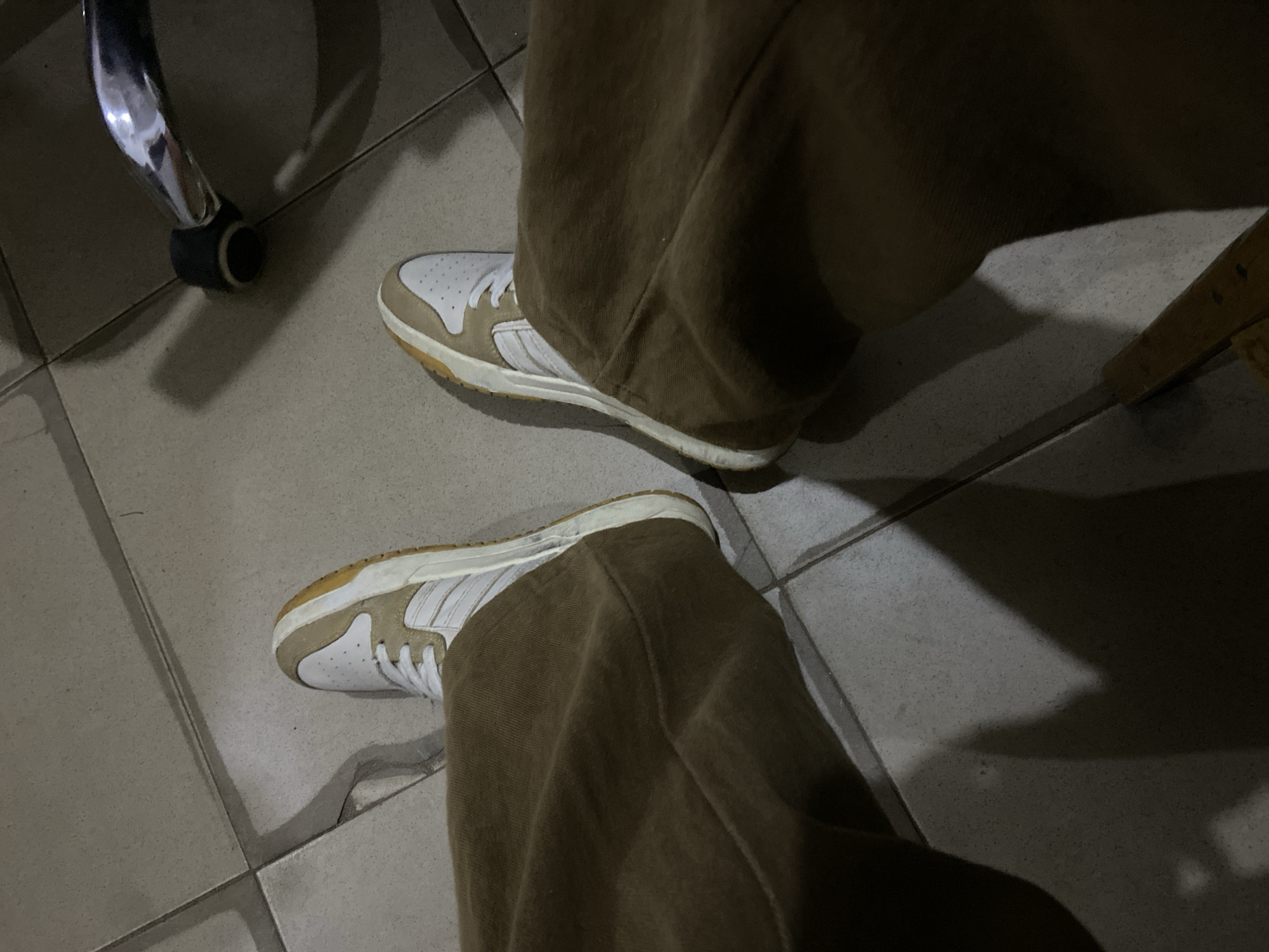 An overhead photograph, taken in dim lighting, captures the lower extremities of an individual standing or seated on a tiled floor in Mararaba, Nigeria. The subject wears loose-fitting, medium-brown trousers and white and tan low-top sneakers. The sneakers feature white uppers with tan suede or synthetic overlays, white laces, and distinct white side stripes, suggestive of an athletic footwear brand. The soles are off-white or light brown, showing signs of wear and minor discoloration. The floor is composed of light grey or beige square tiles, with visible grout lines and some surface debris. In the upper right portion of the frame, the polished chrome leg of a piece of furniture, likely an office chair, is visible, terminating in a black caster or foot. Prominent shadows are cast across the floor by the legs, shoes, and the furniture.