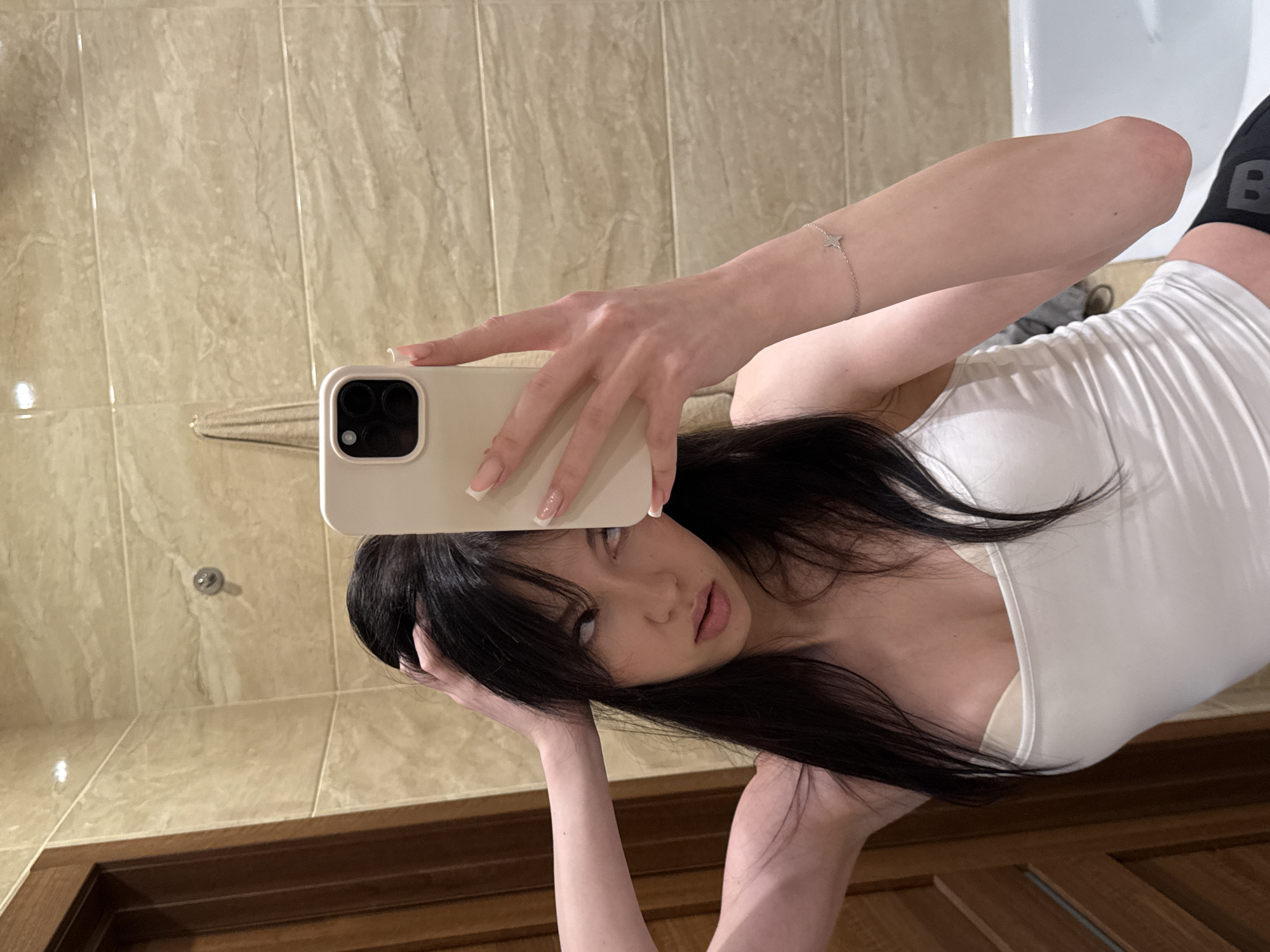 A young woman with long, dark hair is taking a selfie in a bathroom. She is wearing a white tank top and a beige bra. Her right hand is holding a smartphone up to take the picture, and her left hand is in her hair. She is wearing a thin silver bracelet with a star charm on her left wrist. The bathroom has beige tiled walls and a wooden door on the left. A white bathtub is visible on the right side of the image. The lighting suggests an indoor setting, likely a residential bathroom. The image was taken in the city of Khimki, Russia.