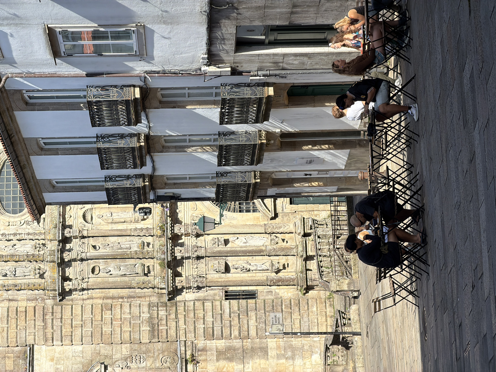 The image depicts an outdoor cafe scene on a sunlit day in Santiago de Compostela, Spain, likely near the Cathedral of Santiago de Compostela. The foreground shows several dark metal tables and chairs set up on a cobblestone plaza. Three groups of people are seated at these tables. On the left, a couple is engaged in conversation, both looking down at their table. In the middle, a man in a black and white striped shirt and light shorts is looking at his phone. To the right, three women are seated, with two appearing to be in conversation and the third looking away.
Behind the cafe, an ornate, aged stone building with intricate carvings and statues dominates the left side of the frame. This building, with its arched doorways and textured stonework, suggests historical architecture. To the right of this, a white building with multiple windows and decorative black metal balconies stands out. The architecture suggests a European city with a rich history. Shadows cast by the buildings and the tables indicate the time of day is likely midday or early afternoon. A street lamp is visible on the stone building, and a street sign is mounted on a pole.