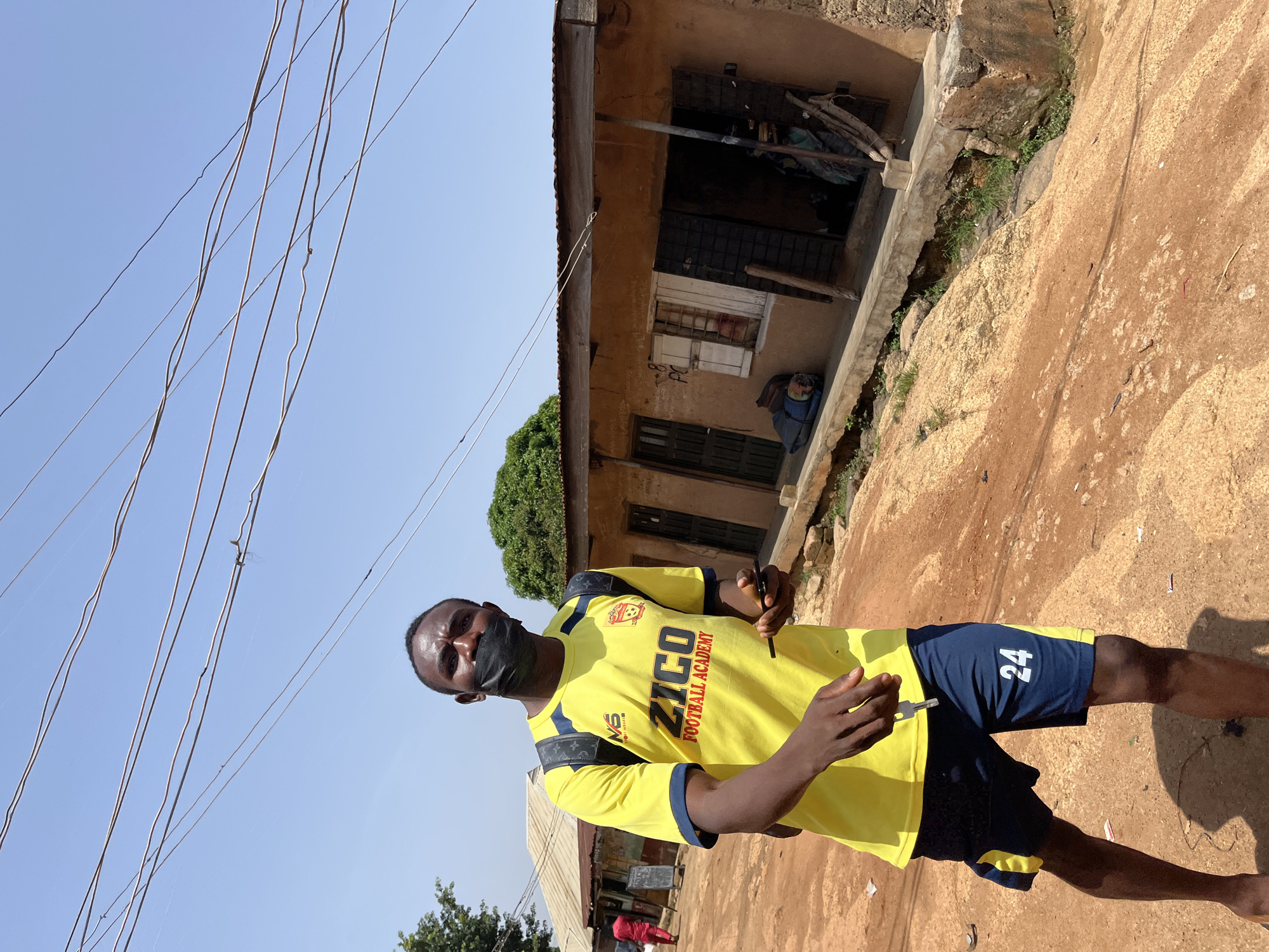 In Jos, Nigeria, a young man wearing a yellow t-shirt with "ZICO FOOTBALL ACADEMY" written on it and dark blue shorts with the number "24" on the left leg, walks on a dirt road. He is wearing a black face mask and carrying a phone in his right hand. A key hangs from his left index finger. He has a black backpack on his shoulders. The background shows a row of low-rise buildings with corrugated iron roofs, typical of some urban areas in Jos. The sky is clear and blue, with power lines running across the frame. The ground is a mixture of dirt and rocks, indicating a dry, possibly dusty environment.