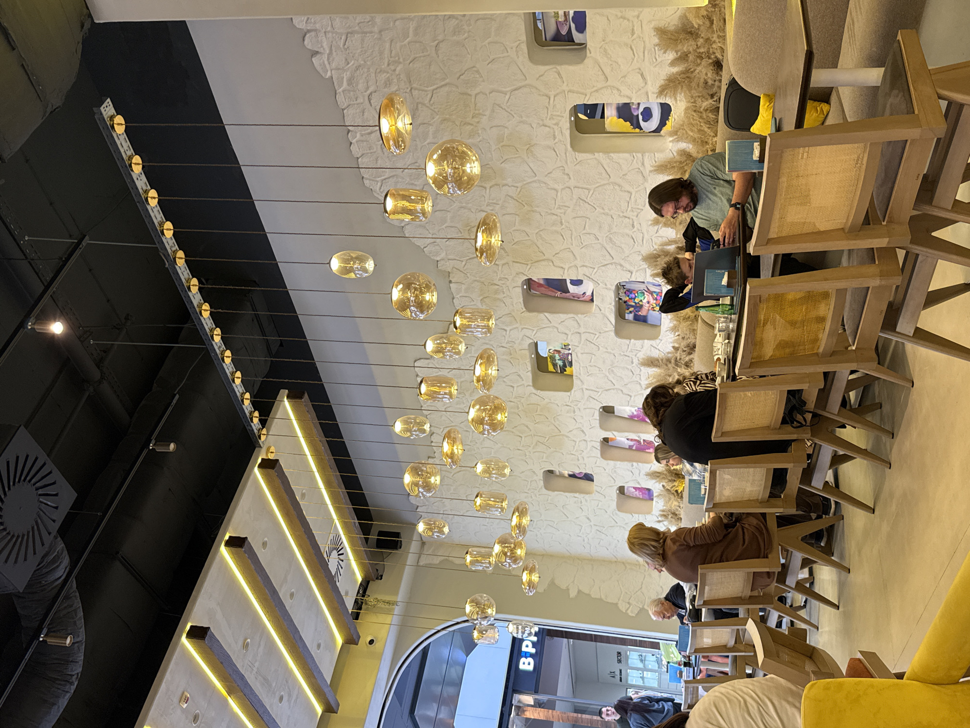 The image depicts the interior of a commercial establishment, potentially a cafe or restaurant, in Belgrade, Serbia. The scene is focused on a row of tables occupied by patrons working on laptops or engaged in conversation. The lighting is provided by a series of hanging pendant lights with decorative bulbs. The walls are textured and adorned with artistic niches containing colorful objects or artwork. The furniture includes light-colored wooden chairs with woven backs. Outside, a glimpse of an exterior space with storefronts and people walking can be seen through a large arched window.