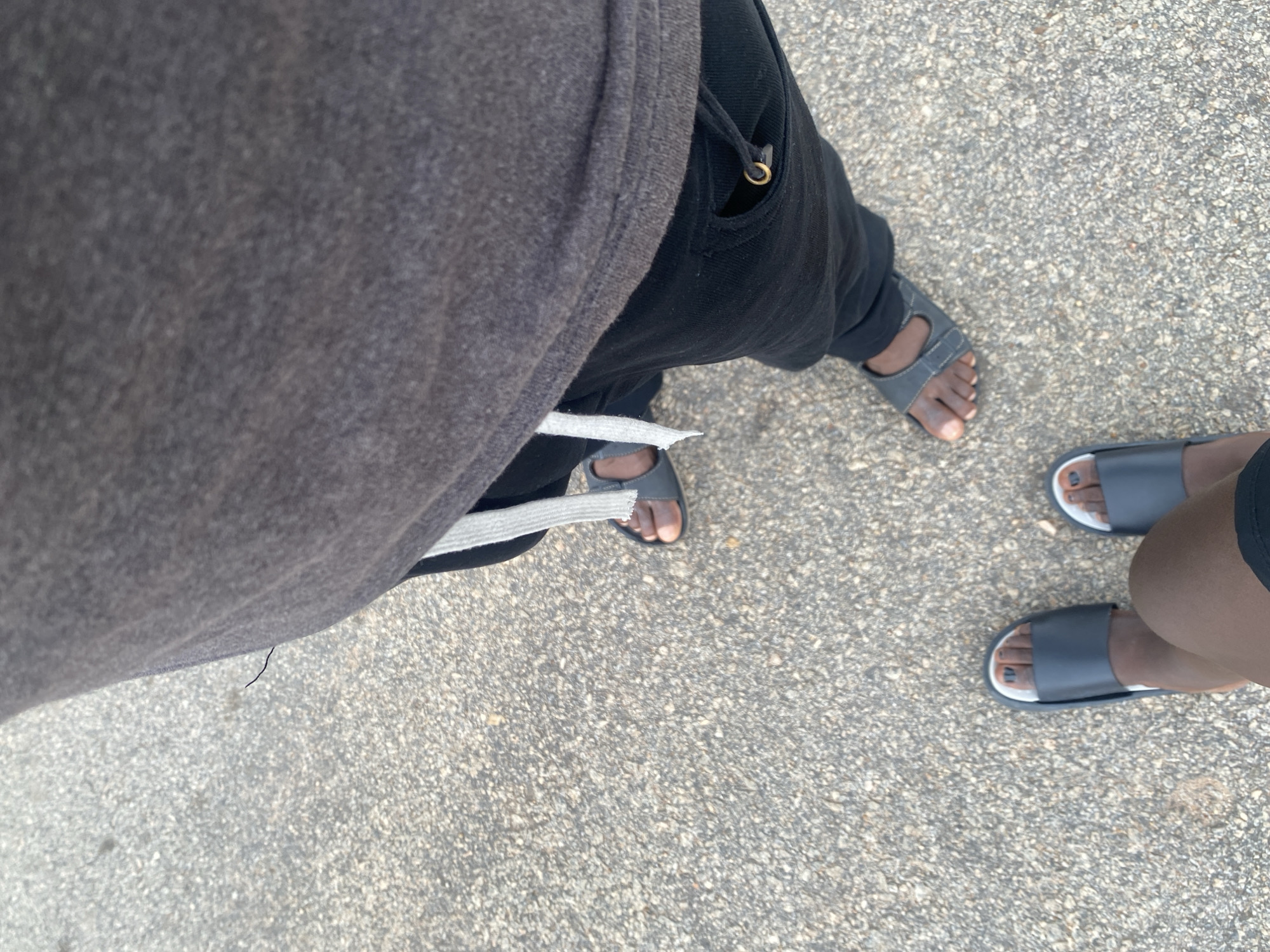 The image shows the legs and feet of two individuals standing on a paved surface, likely outdoors. One person is wearing dark-colored sweatpants and a grey hooded sweatshirt, with one foot visible in a grey sandal. The other person is wearing dark shorts and a pair of dark slide sandals. The ground is a mottled grey and tan, typical of asphalt or concrete, with small pebbles visible. The lighting suggests it is daytime, possibly sunny, but no strong shadows are apparent. The overall scene is casual and appears to be a moment of rest or conversation while walking. The image is taken from a high angle looking down. There is no visible text in the image. The context provided is Dumnga, Nigeria.