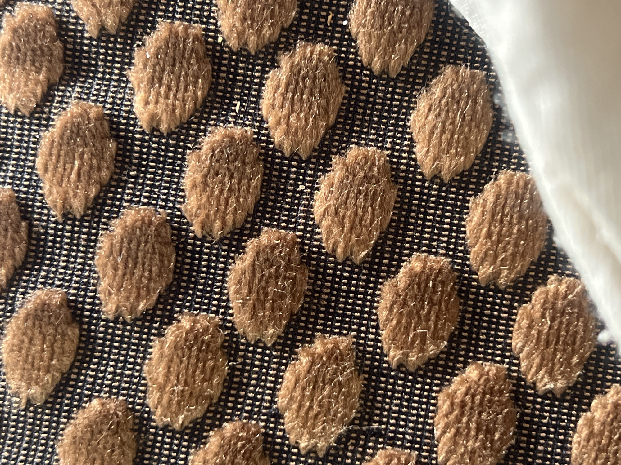 The image displays a detailed, close-up view of a textured fabric. Numerous distinct, raised, and irregularly shaped fuzzy brown elements are arranged in an offset grid pattern across the frame. These elements appear soft and fibrous, with individual strands reflecting light, giving them a slightly shimmering or textured appearance. They are set against a darker, likely navy blue or black, woven background fabric. This background exhibits a fine, consistent grid-like weave, with subtle lighter-toned threads visible within its structure. The lighting accentuates the three-dimensional quality of the raised brown components. No people, discernible actions, or environmental details are present in the image, and there are no visual cues to suggest a specific location, including the city of Casablanca, Morocco. The scene is a forensic examination of the textile's pattern, texture, and material properties.