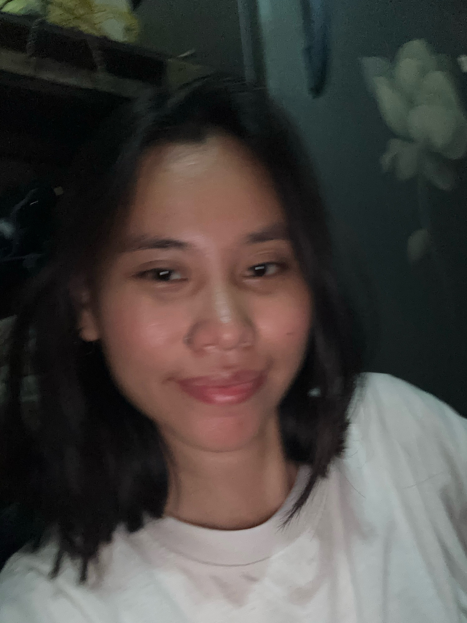 The image shows a close-up, slightly blurred selfie of a young woman with dark hair and fair skin. She is wearing a white t-shirt. Her expression is neutral to slightly smiling, with her lips slightly parted. The background is dark and out of focus, with hints of what appears to be a patterned wallpaper or fabric on the right side, possibly depicting floral motifs. The lighting is dim. The scene is indoors, likely in a residential setting. There are no specific city landmarks or environmental cues that would allow for definitive identification of East Jakarta, Indonesia.
