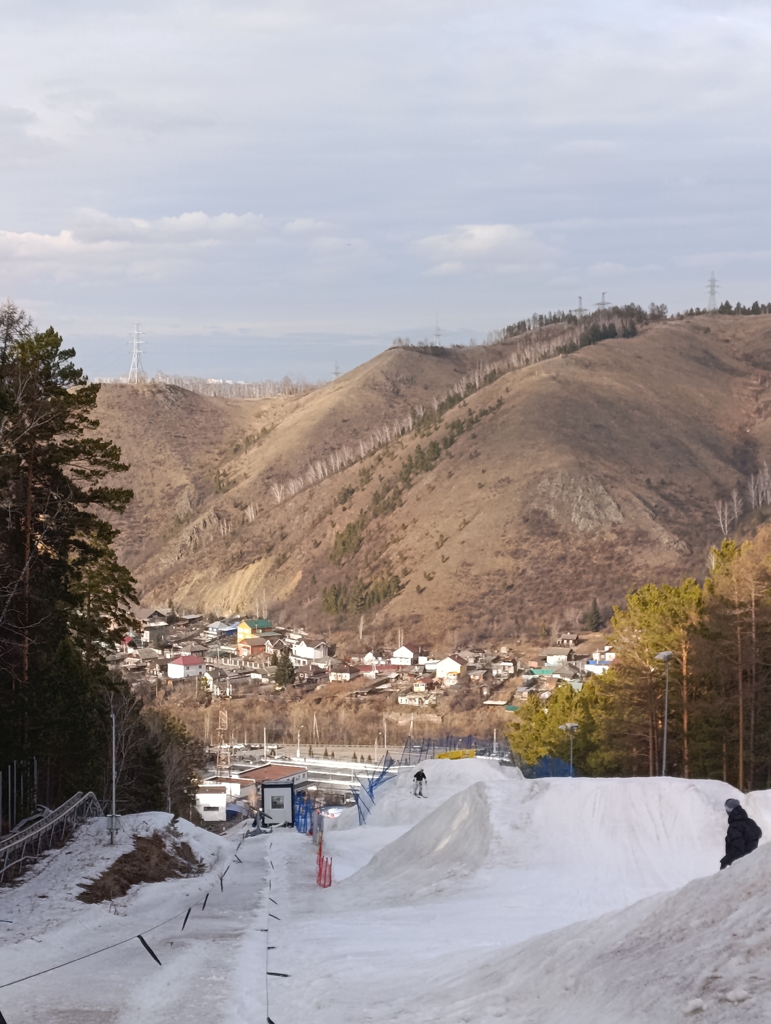 The image shows a snow park in Krasnoyarsk, Russia, with a view of the city and surrounding hills. In the foreground, there's a snow-covered area with ramps and jumps, presumably for skiing or snowboarding. At least two people are visible: one in the middle distance on skis or a snowboard, and another individual standing on the snow near the right edge of the frame. A small building or structure, perhaps related to the snow park facility, is visible at the base of the snow-covered area. In the background, a densely built area with houses and buildings is nestled at the foot of hills. The hills are covered with sparse trees and what appear to be power lines running along their crests. The sky is overcast with muted light. The scene suggests a cold climate and the use of the location for winter sports activities.