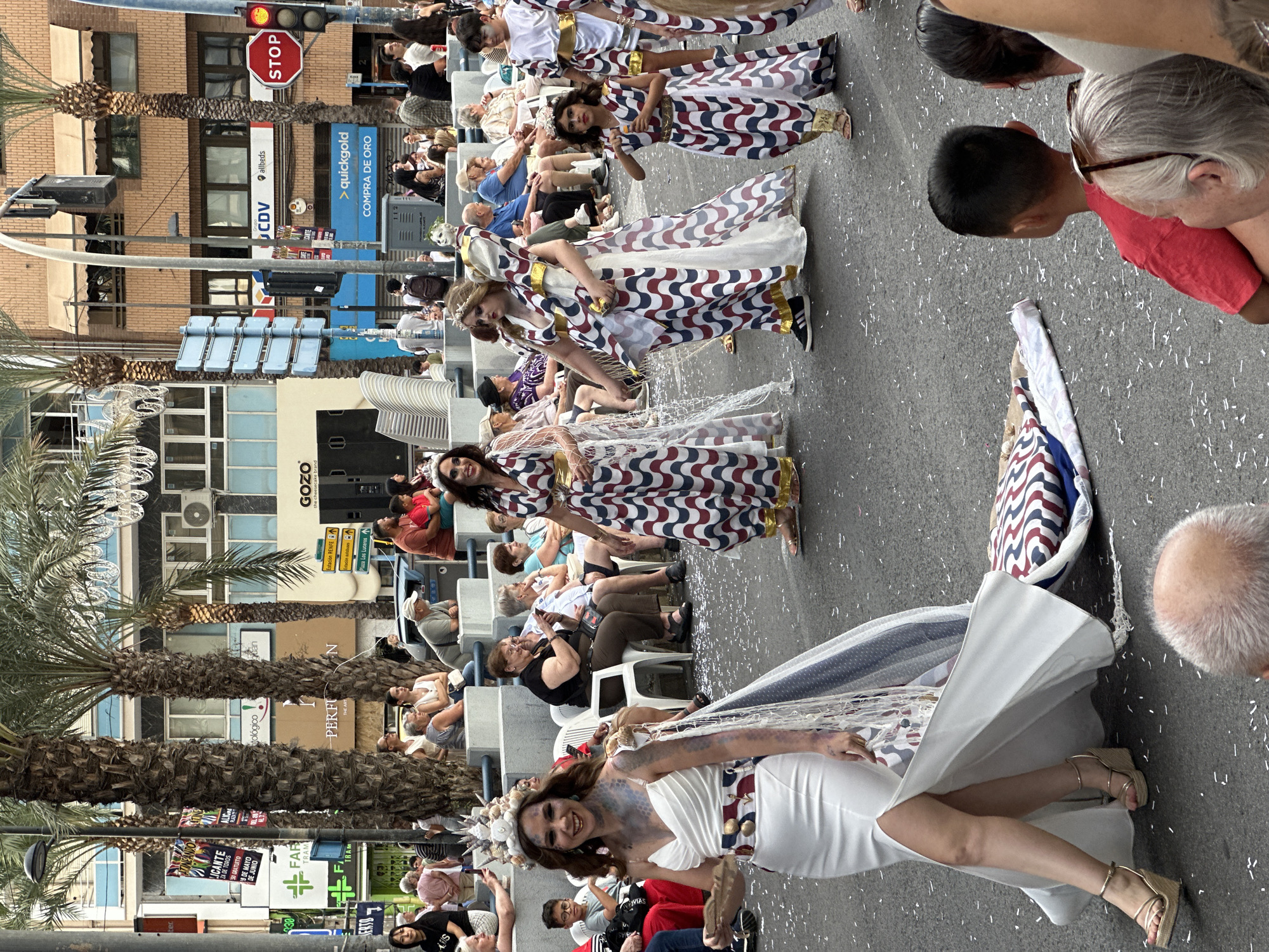 This image depicts a festive street parade in Alicante, Spain, under daylight conditions. The scene is dominated by several parade participants in elaborate costumes, walking down an asphalt street strewn with white confetti.
In the immediate foreground, a woman with dark hair, fair skin, and blue-green face paint on her left cheek smiles directly at the viewer. She is wearing a strapless, floor-length white gown with a high left-leg slit, revealing platform wedge sandals with ankle straps. Her head is adorned with a large, intricate headdress made of what appears to be shells and coral, resembling a sea-themed motif. A decorative cape or train, partially white and partially patterned with blue dots and a red, white, and blue wavy design, trails behind her, with its end resting on the street. Her right arm is extended forward, holding a brown, fan-like object. A decorative, possibly scaled or textured element, extends from her left shoulder down her arm.
Behind her, to the right, another woman with dark hair walks in a long dress featuring a red, white, and blue wavy pattern and a golden belt. Her dress also appears to have a trailing element. Further back and to her right, a third woman, similarly dressed in the wavy patterned gown with a golden belt and a crown, walks ahead. To her right, a young girl, also in a matching wavy patterned dress with a golden belt, walks while holding a small, clear object in her hand. A young boy in a similar patterned dress is partially visible behind the girl.
On both sides of the street, spectators observe the parade. Many are seated on white plastic chairs or ledges behind metal barriers. They are diverse in age and attire, dressed in casual street clothes. In the far foreground, the top of a man's bald head and a young boy's dark-haired head are visible, watching the parade.
The background features multiple urban buildings, some with awnings and balconies. Several palm trees line the street. Visible signs include "ALICANTE" on a banner, "GOZO the cheesecake brand," "quickgold COMPRA DE ORO" (Quickgold Buy Gold), "FARMACIA TRAM," and "Estación RENFE" (RENFE Station) directions. A red stop light is visible on the right side of the street, along with street lamps and overhead decorative lighting. The sky is partially visible at the top, suggesting an overcast or bright day.