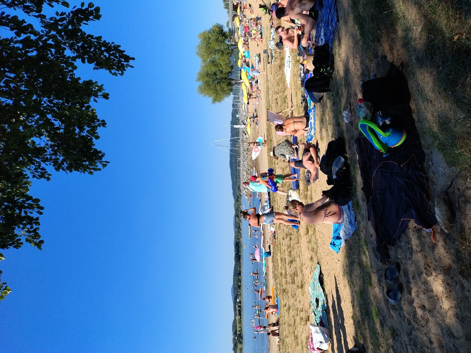 This is a full-shot, eye-level, outdoor photograph of a crowded beach scene on a sunny day. The scene appears to be a lake beach in Radslav, Czechia, given the context of the provided crops. The sky is a clear, bright blue. In the foreground, several people are sunbathing on towels and blankets spread on the sandy beach. Some are lying down, while others are sitting. There are various items scattered on the sand, including towels, bags, and a pair of flip-flops.
In the mid-ground, the sandy beach extends to the edge of a body of water, likely a lake. Several people are in the water, some on paddleboards and others swimming. Boats and sailboats are visible on the water in the distance. More people are scattered along the shoreline, some sitting or standing. There are numerous yellow umbrellas providing shade on the beach.
In the background, a tree-lined landscape is visible across the lake, with hills in the far distance. The overall atmosphere is one of a busy summer day at a recreational area.