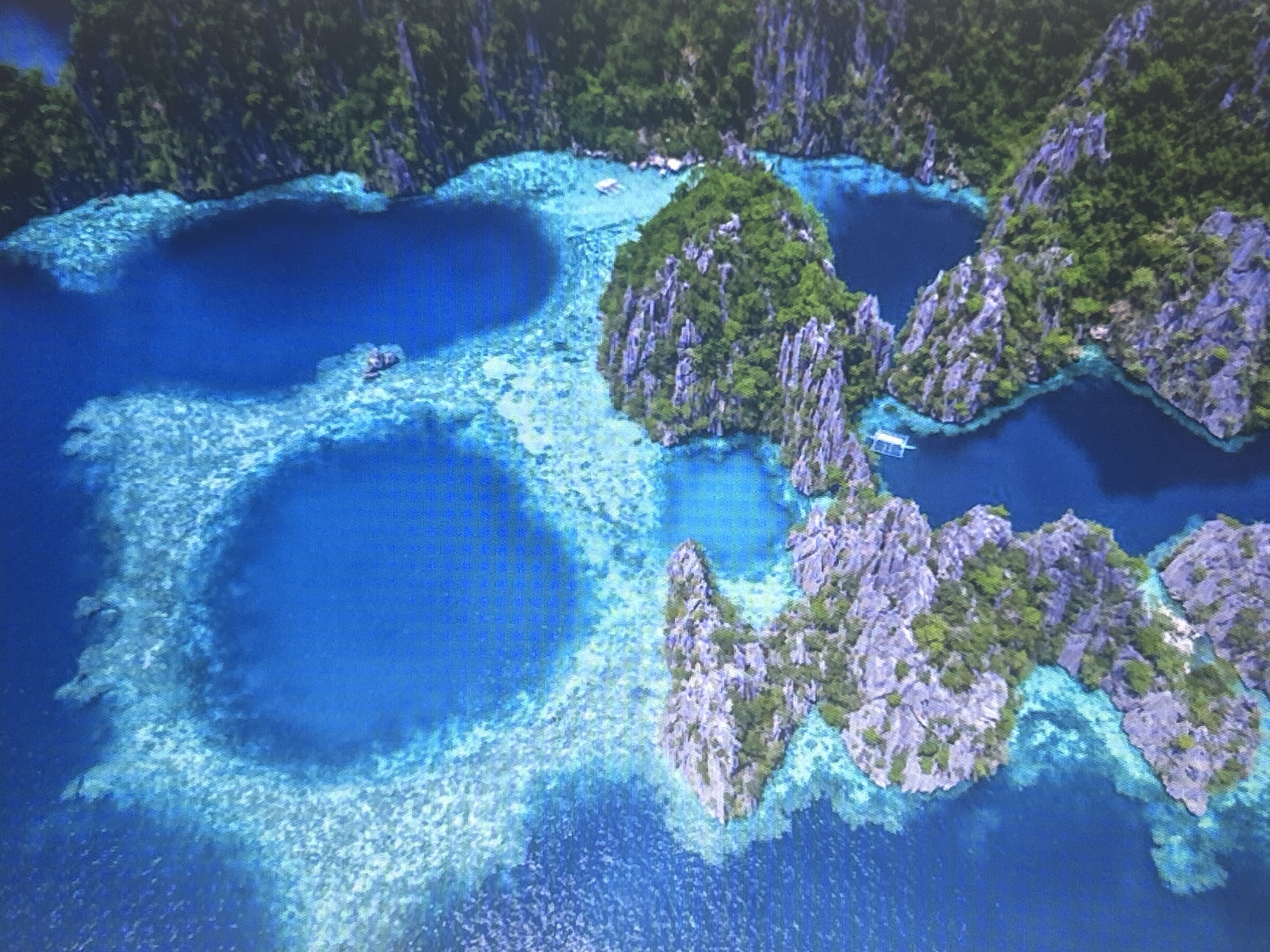 This is an aerial photograph of a tropical island landscape featuring steep, verdant limestone cliffs surrounding intensely blue lagoons. The water is exceptionally clear, revealing submerged coral reefs in lighter shades of turquoise and white. There are a few small boats visible on the water. The image captures a pristine natural environment.
This image is not of Sofia, Bulgaria. It depicts a tropical island, likely in Southeast Asia, given the characteristic karst topography and clear blue waters associated with destinations like Palawan in the Philippines or Halong Bay in Vietnam. Sofia is a landlocked city in Bulgaria and does not have coastal features or this type of tropical island scenery.