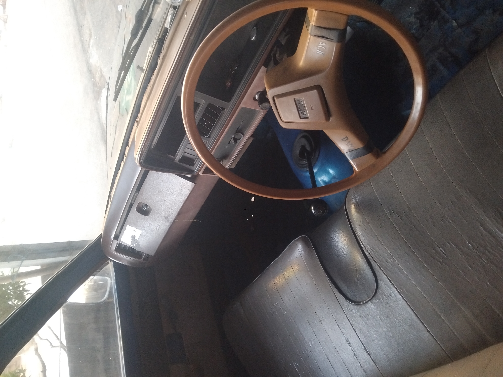 The image shows the interior of a car, viewed from the passenger seat, looking towards the driver's side. The car appears to be parked outdoors, possibly in a residential area. The interior is dated, with a brown dashboard and a wood-grain steering wheel. The seats are upholstered in dark gray vinyl. The dashboard has a rectangular instrument cluster with various gauges and controls, as well as air vents. The steering wheel has a Chevrolet logo in the center. The floor is covered in a blue mat. The exterior of the car is visible through the windows, including a tree and a concrete wall. The lighting suggests it is daytime, possibly overcast or late afternoon. No people are visible in the image. The car is located in the city of Tempeh, Indonesia.