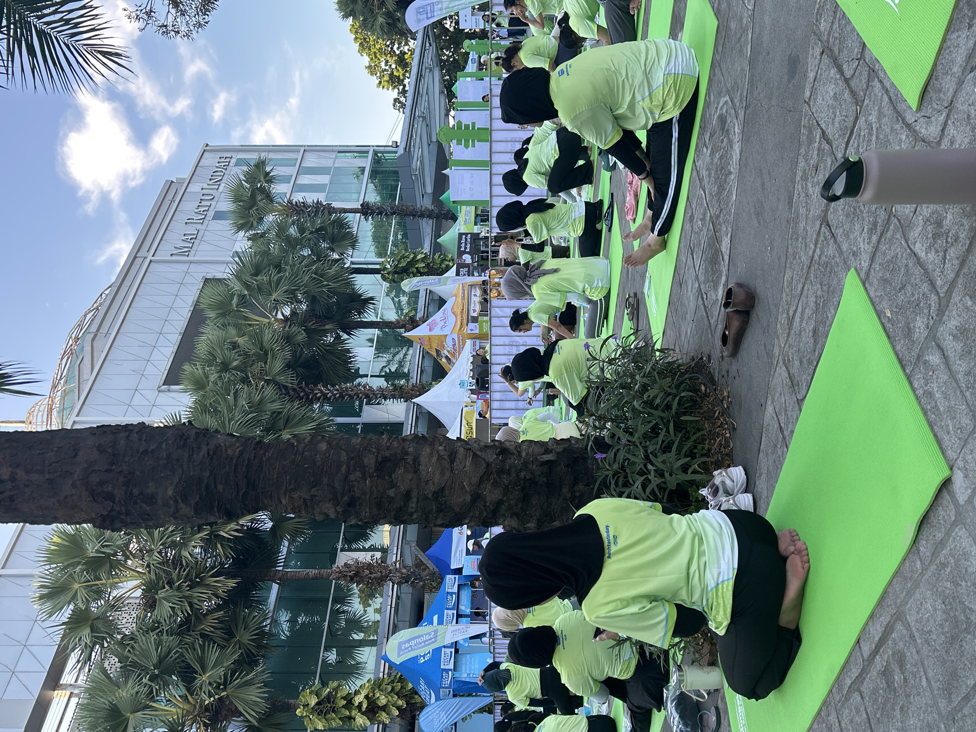 The image depicts an outdoor yoga session taking place in front of the Mal Ratu Indah shopping mall in Makassar, Indonesia. A group of individuals, mostly women wearing lime green t-shirts and headscarves, are seated or in various yoga poses on bright green yoga mats laid out on a paved surface. The backdrop features the modern architecture of the mall with its glass facade and signage, along with palm trees and blue sky with clouds. Several tents and banners are visible behind the participants, suggesting an organized event. Objects like water bottles, shoes, and bags are scattered among the yoga mats. The scene is captured from a slightly elevated perspective, focusing on the participants engaged in their activity.