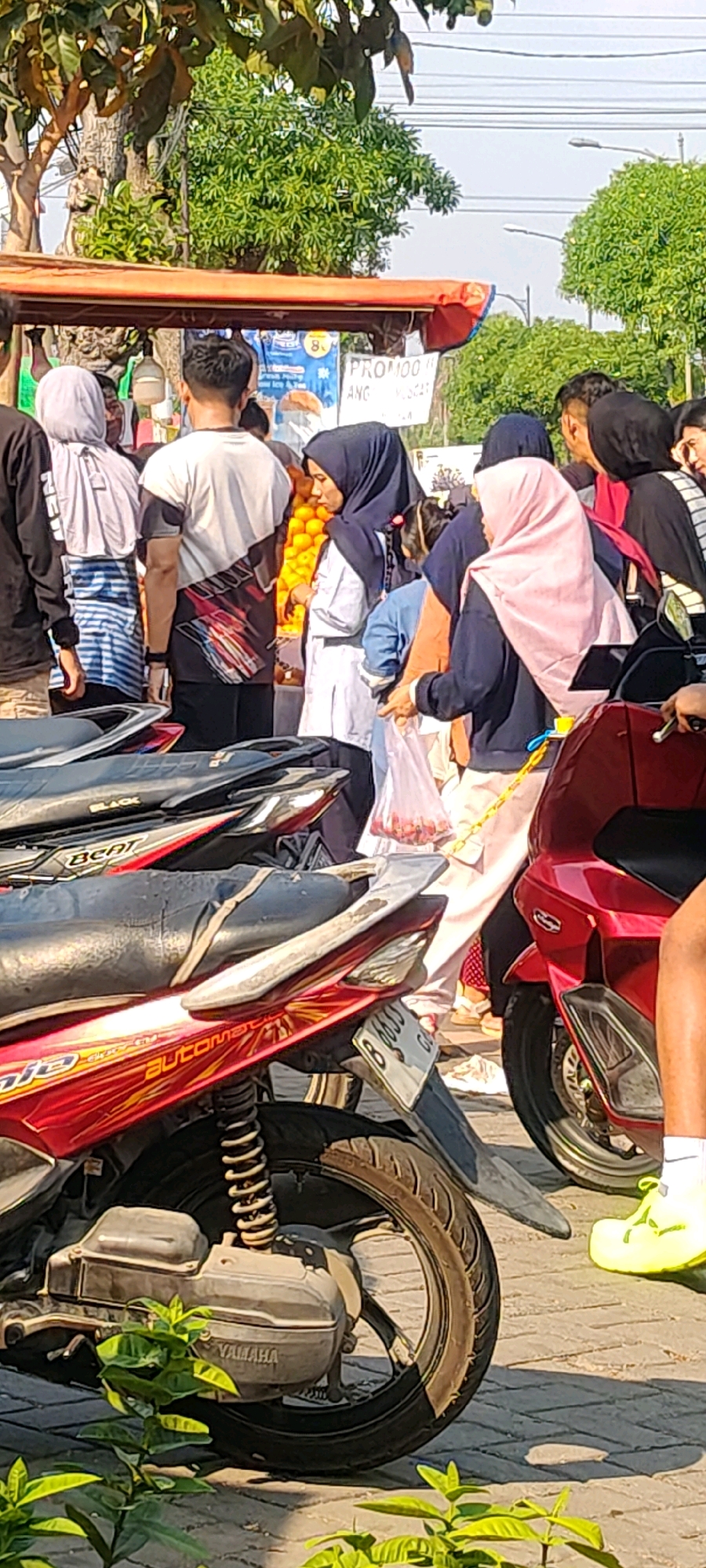 In Cibatu, Indonesia, a street scene unfolds with a group of individuals gathered around a stall, possibly a fruit stand given the visible oranges. Several motorcycles are parked, taking up a significant portion of the foreground. A red motorcycle on the right features a visible license plate, and a person's legs clad in bright yellow sneakers and white socks are visible near it. The people in the background are diverse, with some wearing headscarves, suggesting a mixed-gender crowd. One person is holding a clear plastic bag filled with small, red items, possibly fruits or candies. A sign with the word "PROMO" is partially visible behind the people. The environment suggests an outdoor market or roadside vending area, with trees and power lines in the background, indicating an urban or semi-urban setting. The lighting suggests it is daytime.