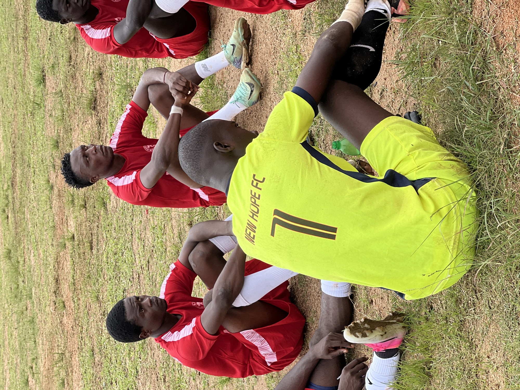 A group of young men in soccer uniforms are sitting on a dirt and grass field. The men are wearing red jerseys with white stripes on the sleeves and red shorts. One man in the foreground is wearing a bright yellow jersey with the words "NEW HOPE FC" and the number "7" on the back. The men are sitting with their legs crossed or extended, and some are looking away from the camera. The environment appears to be an outdoor sports field with dry, brown earth and patches of green grass. The lighting suggests it is daytime. This scene could be taking place in Jos, Nigeria, a city known for its soccer culture.