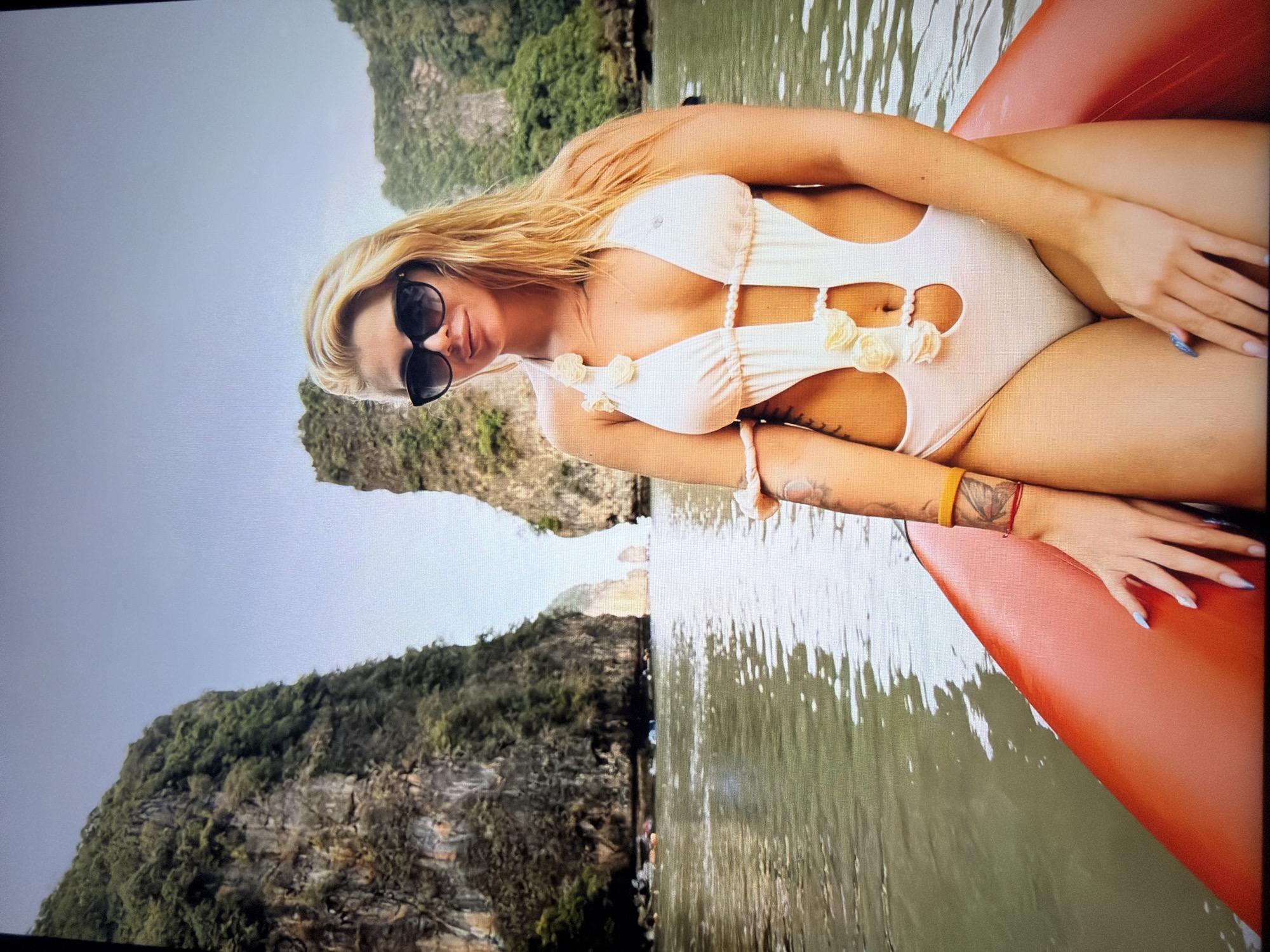 The image, oriented horizontally, depicts a young woman with long, flowing blonde hair, wearing black sunglasses and a light-colored monokini with cut-out designs and white floral appliqués. Her fingernails are painted light blue, and she wears a yellow/gold band bracelet and a red string bracelet on her right wrist, along with a tattoo of what appears to be a flower on her forearm.
She is seated in what looks like a reddish-orange inflatable raft or kayak, positioned on a body of calm, reflective water. The woman is looking directly at the viewer with a neutral, slightly smiling expression. The background features striking natural scenery: towering, tree-covered rocky cliffs or limestone karsts rise prominently from the water, suggesting a bay or large inlet. The sky is a uniform, overcast grey.
The scene captures a leisurely moment, likely a vacation or recreational activity such as kayaking or exploring, in a tropical or subtropical natural setting during daylight hours. The specific geological formations strongly suggest a location in Southeast Asia. No text is visible in the image.