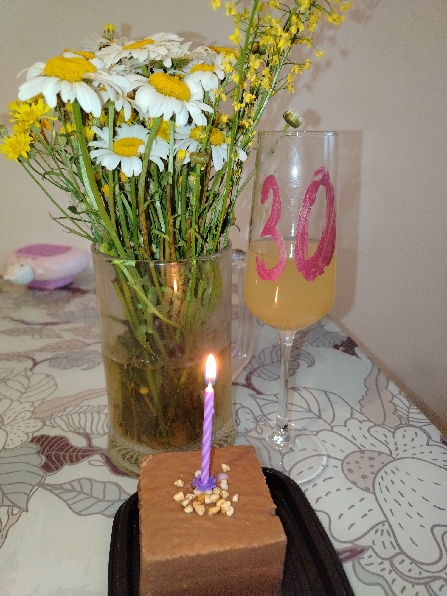 The image depicts a close-up, indoor scene, likely a celebration, with a birthday cake and drink. A small chocolate cake with a single purple candle and what appear to be nuts or sprinkles on top is in the foreground. To the left of the cake is a glass vase filled with a bouquet of white daisies and yellow flowers. A lit purple birthday candle is positioned between the cake and the vase. To the right of the vase is a champagne flute containing a yellow beverage, with the number "30" painted in pink on the outside. The items are arranged on a table covered with a patterned cloth. In the background, out of focus, is a pink object with a white nose, resembling a decorative item. The wall in the background is a plain, light color. No specific location within Noviy, Russia can be identified from the image.