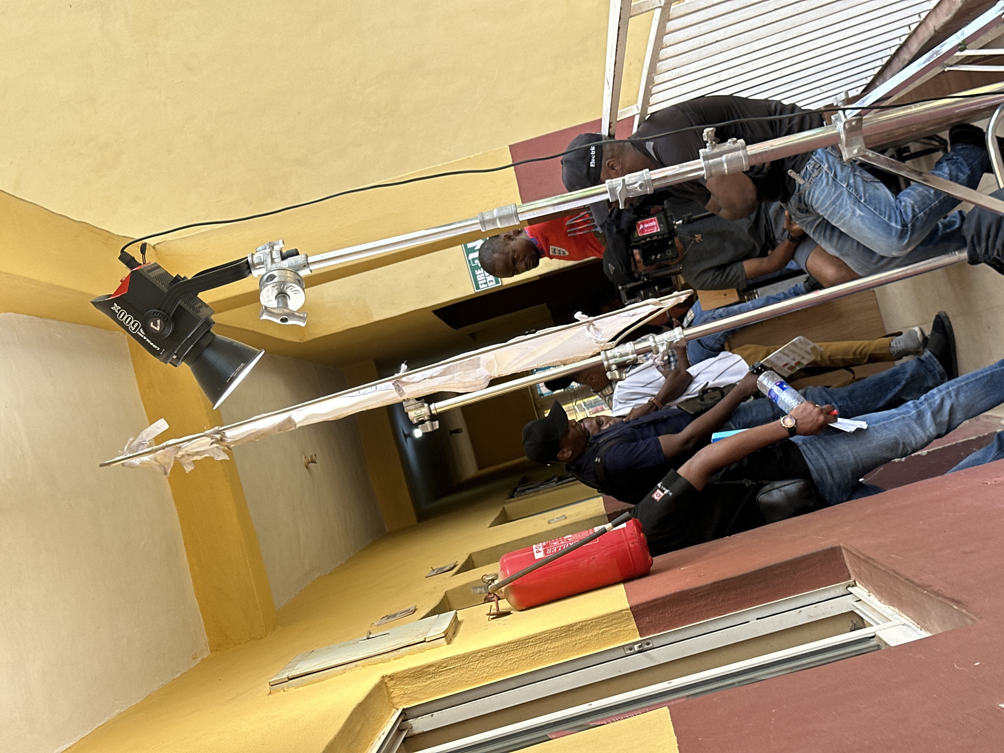 The image shows an indoor scene, seemingly on a film set, with crew members working. A professional light fixture, branded "Aputure 600x", is suspended from a silver boom arm. Two men are positioned on scaffolding, one operating a professional video camera, identifiable by its red markings. A red fire extinguisher lies on a maroon and yellow wall. Additional crew members are present, one holding a water bottle. The environment suggests an urban setting, potentially a building interior in Lagos, Nigeria, given the architectural style and the presence of equipment typically used in film production.