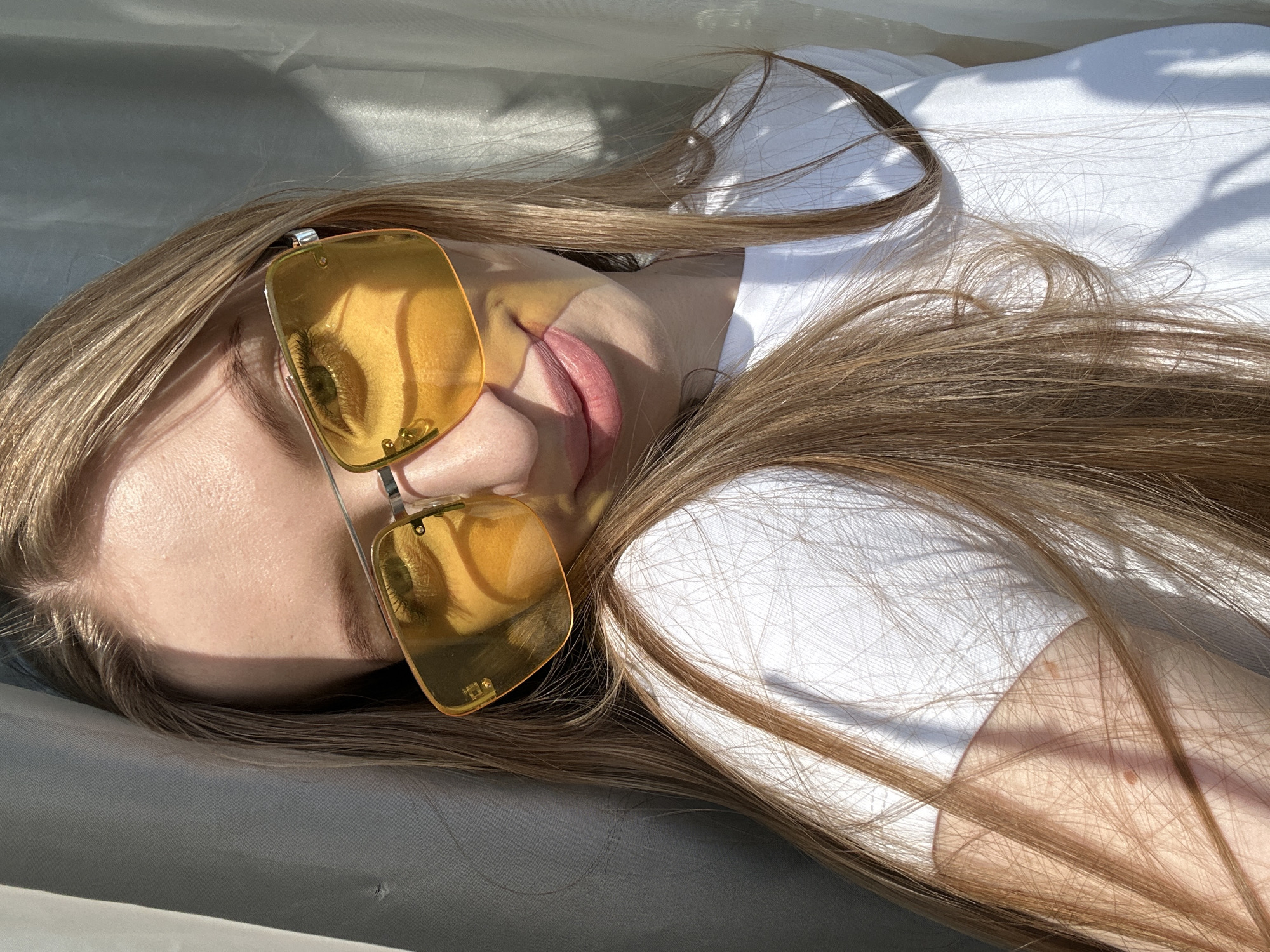A young, fair-skinned woman with long, light brown hair is lying down, wearing a white t-shirt and gold-tinted rectangular sunglasses. The sunglasses reflect her eye. She appears to be outdoors, possibly in Chaikovsky, Russia, based on contextual knowledge. The scene is bathed in sunlight, casting shadows on the fabric she is lying on.