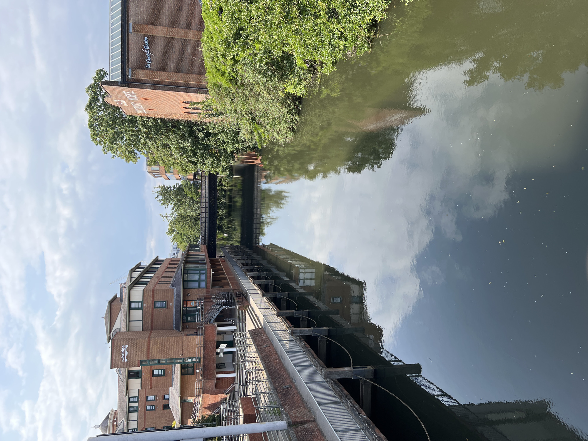 The image shows a view of the River Wey in Guildford, United Kingdom. The river flows between two brick buildings. On the left is a building with the word "thebridge" on it, which appears to be a modern office building or residential complex. It has multiple stories, brickwork, and large windows. A walkway with metal railings runs along the riverbank in front of this building. On the right is an older brick building with arched windows and the text "1913 ELECTRICITY WORKS" and "The Electric Theatre" visible on its facade. This building is covered in ivy in places, and dense green foliage borders the river. A footbridge crosses the river in the background, connecting the two sides. The water reflects the sky and the surrounding buildings and trees. The sky is partly cloudy. The scene appears calm and tranquil, with no people clearly visible in the foreground.