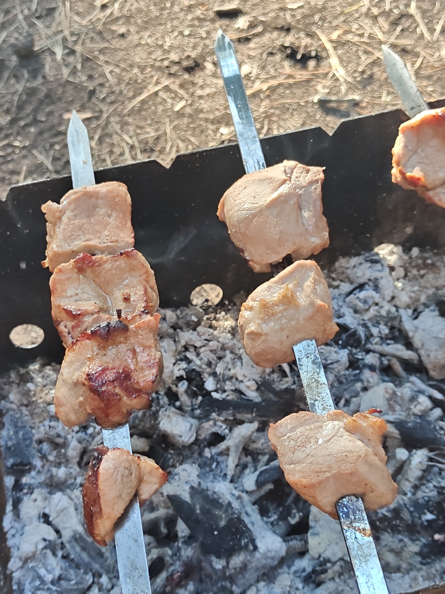 This image shows a close-up, overhead view of several skewers of meat cooking over hot coals in a metal brazier. The meat appears to be pork, cut into cubes and threaded onto metal skewers. Some pieces are showing signs of browning and slight charring from the heat. The skewers are positioned at different angles within the brazier, which contains a bed of grey ash and glowing embers. The background is a textured, brown surface, possibly ground or dry vegetation, with some sunlight glinting off the metal skewers. The scene is set outdoors, with the natural light suggesting a daytime setting, and the overall context points to a barbecue or outdoor cooking event. No specific landmarks or identifying features of Корнеев ауылдық округі, Kazakhstan, are visible in the image.