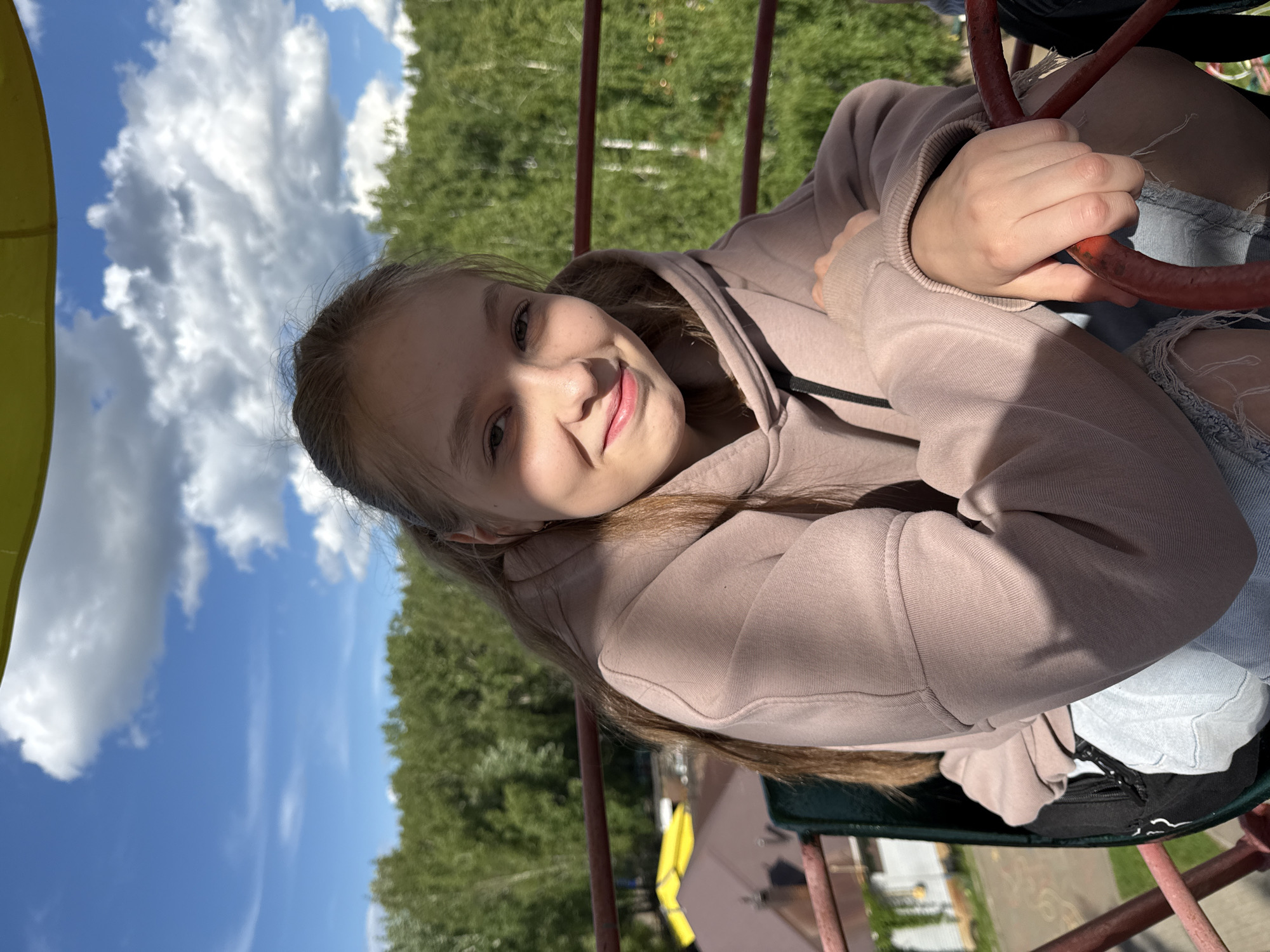 The image shows a young girl in Chaikovsky, Russia, seated on a ride that resembles a small, manually operated ferris wheel. She is wearing a light brown hoodie and holding onto a red metal bar of the ride. She is smiling at the camera.
The background shows a blue sky with white clouds and green trees. There is a partial view of yellow playground equipment and other structures, like buildings, are visible below.