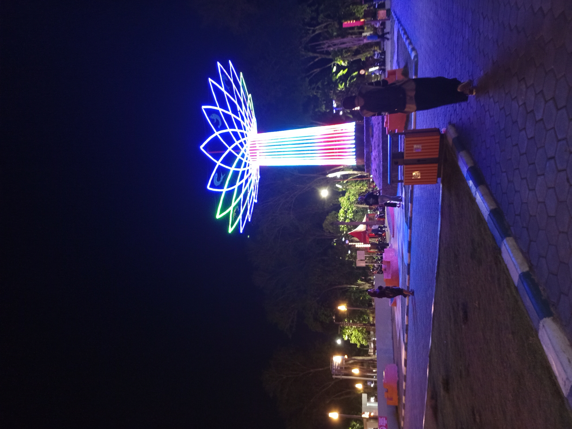 At night in Sidoarjo, Indonesia, a large, illuminated tree-like sculpture dominates the scene. The sculpture, composed of bright neon lights in shades of blue, green, red, and white, stands tall in a park-like setting. The upper portion of the sculpture features a lotus flower design, outlined in blue and green neon. The trunk is illuminated with vertical stripes of red, white, and blue.
In the foreground, a person wearing a dark hijab and a long dark skirt is walking away from the camera, casting a long shadow on the hexagonal paved walkway. Beside the walkway, a person in dark clothing is visible walking towards the sculpture. In the mid-ground, several other people are seen walking and standing in the vicinity of the sculpture. The background shows a line of trees and streetlights, suggesting a public space or park within the city. The overall atmosphere is one of a vibrant urban night scene, likely a tourist attraction or a public event space in Sidoarjo.