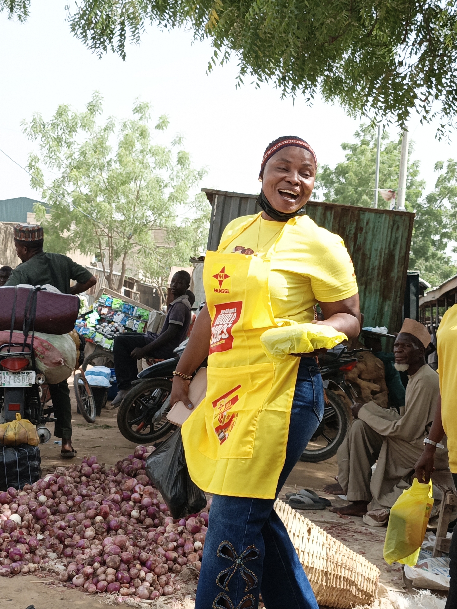 This vibrant image captures a bustling outdoor market scene in Kawaji, Nigeria, under bright, clear daylight. The central figure is a cheerful woman, smiling broadly with an open mouth, exuding enthusiasm. She wears a bright yellow t-shirt, blue jeans with distinctive embroidered patterns, and a prominent yellow apron. The apron clearly displays the "MAGGI" logo and "DOUBLE STAR" branding, suggesting she is engaged in a brand promotion or sales activity. A black face mask is pulled down to her chin, and a colorful patterned headband adorns her head. She holds a yellow plastic bag in one hand and a phone in the other.
The market environment is characterized by active commerce. In the immediate foreground, a large, sprawling pile of red onions dominates the ground, indicating the sale of fresh produce. Surrounding the woman are various market-goers and vendors. To her left, a man in a dark green shirt stands beside a motorcycle heavily laden with large sacks and luggage. Another man is seated on a separate motorcycle, which is piled high with numerous small, brightly packaged items. On the right, an older man with a white beard and a traditional cap sits, observing the lively scene. Partially visible on the far right is another individual, also dressed in a yellow top, possibly part of the same promotional team.
The background reveals typical market infrastructure, including simple structures made of corrugated metal and verdant trees under a bright sky. A visible motorcycle license plate, "KANO JJJ 5500", firmly places the scene within the Kano State of Nigeria, consistent with the Kawaji location context. The overall impression is one of a busy, community-focused market bustling with daily life and commercial activity.